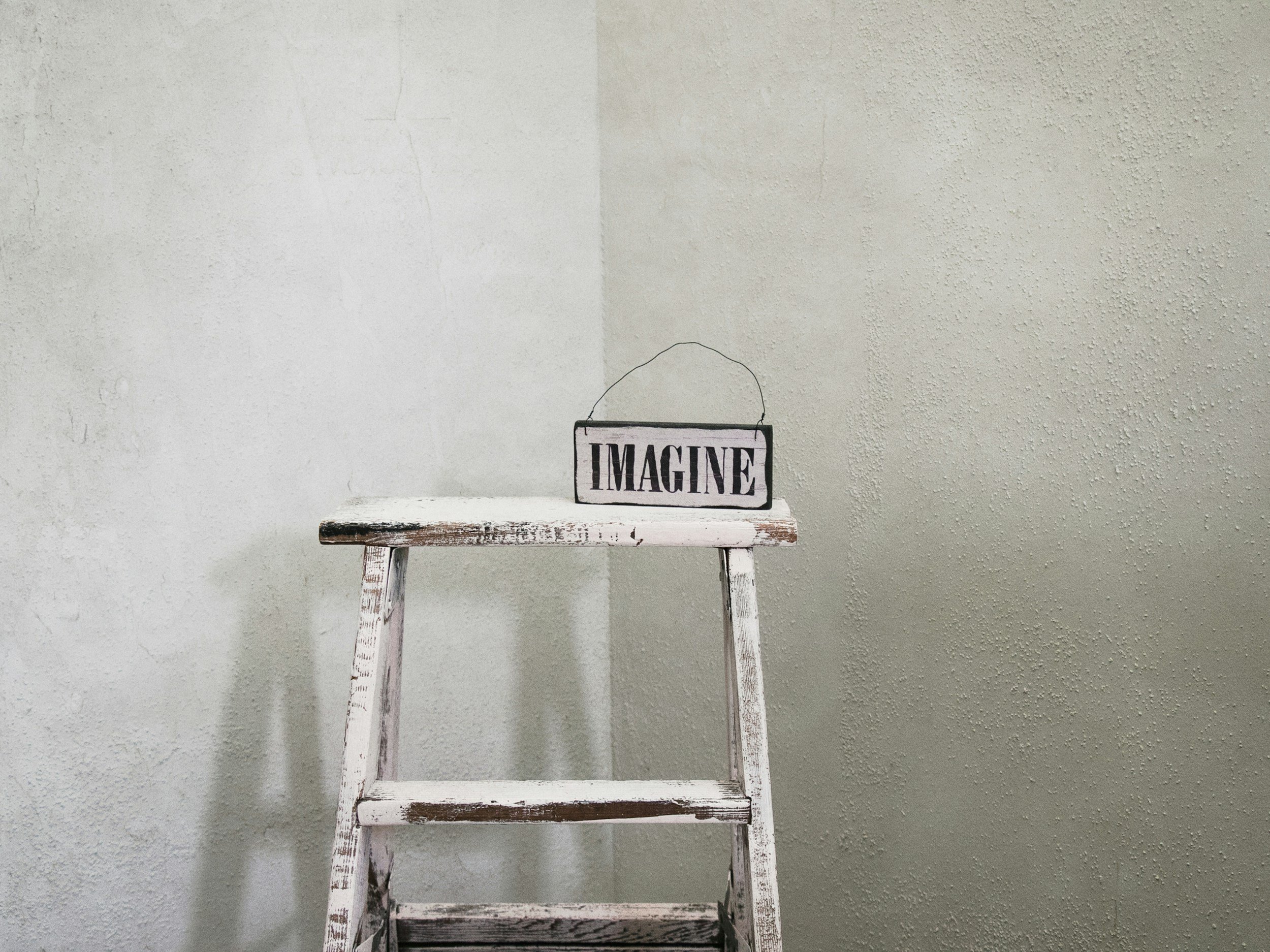 A distressed white wooden ladder stands against a textured wall painted half white and half beige. Sitting ontop of the ladder is a small rectangular sign with the word 'IMAGINE' printed in bold black letters.