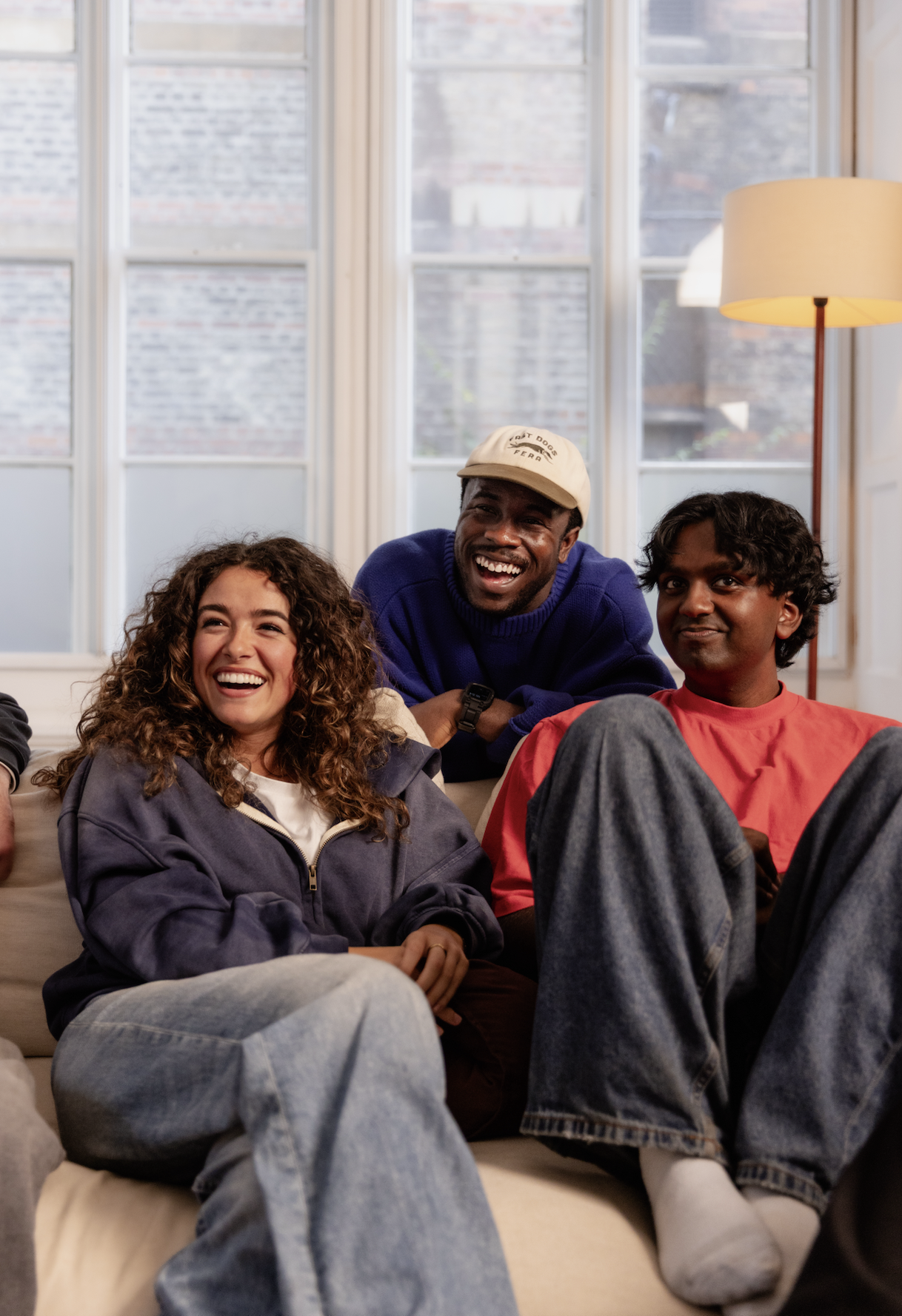 Three friends sitting on a couch, smiling and laughing together, with a bright window and a tall floor lamp in the background.