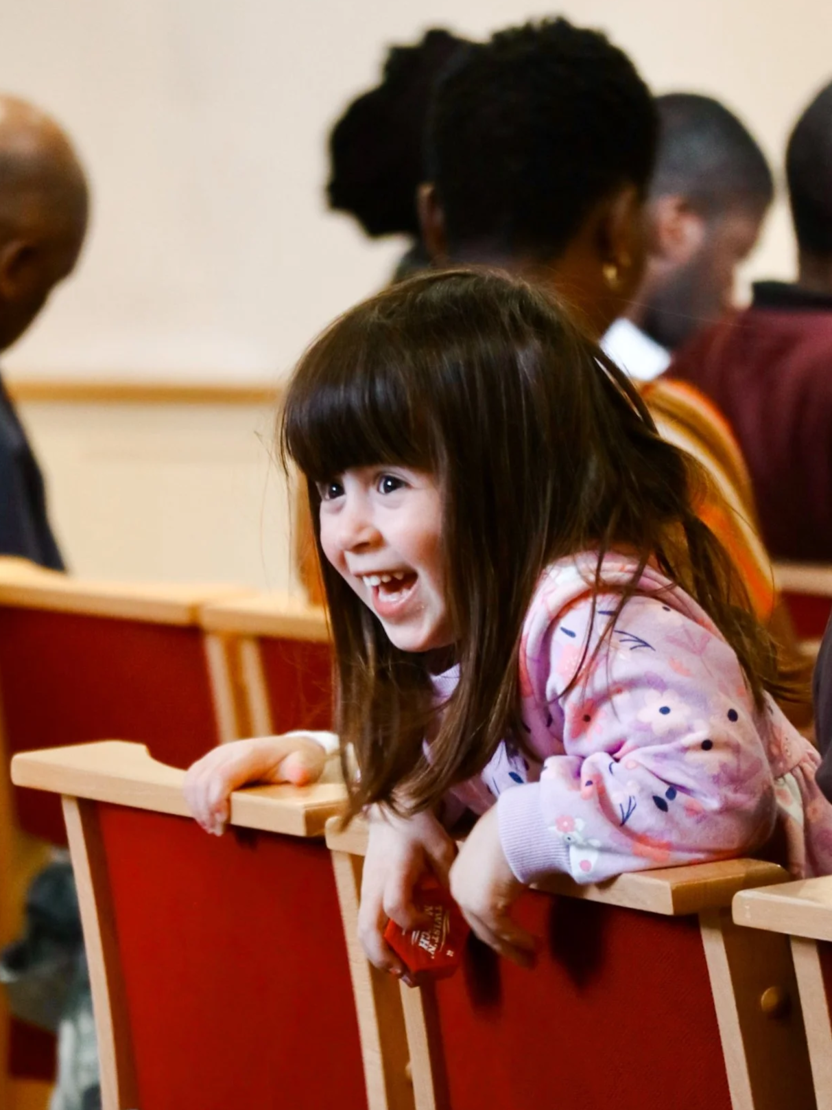 A young girl with long brown hair and a pink patterned hoodie leaning over a wooden pew, smiling and holding a snack, in a church or auditorium setting with other people seated in the background.