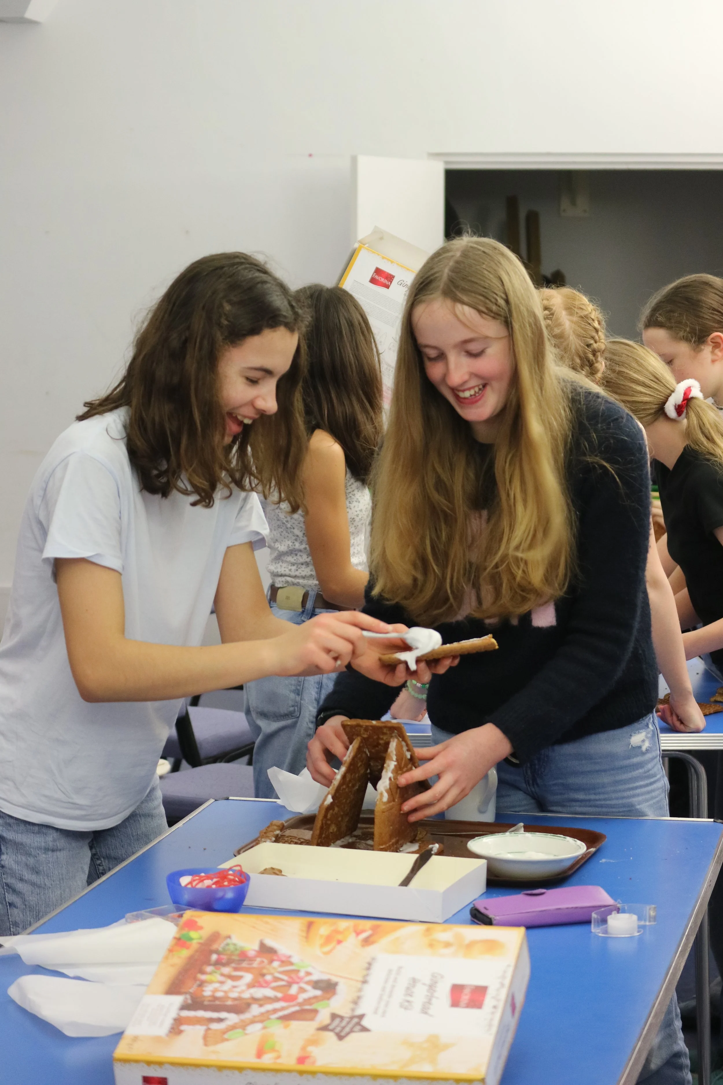 Group of young girls decorating gingerbread houses at a table, smiling and having fun indoors.