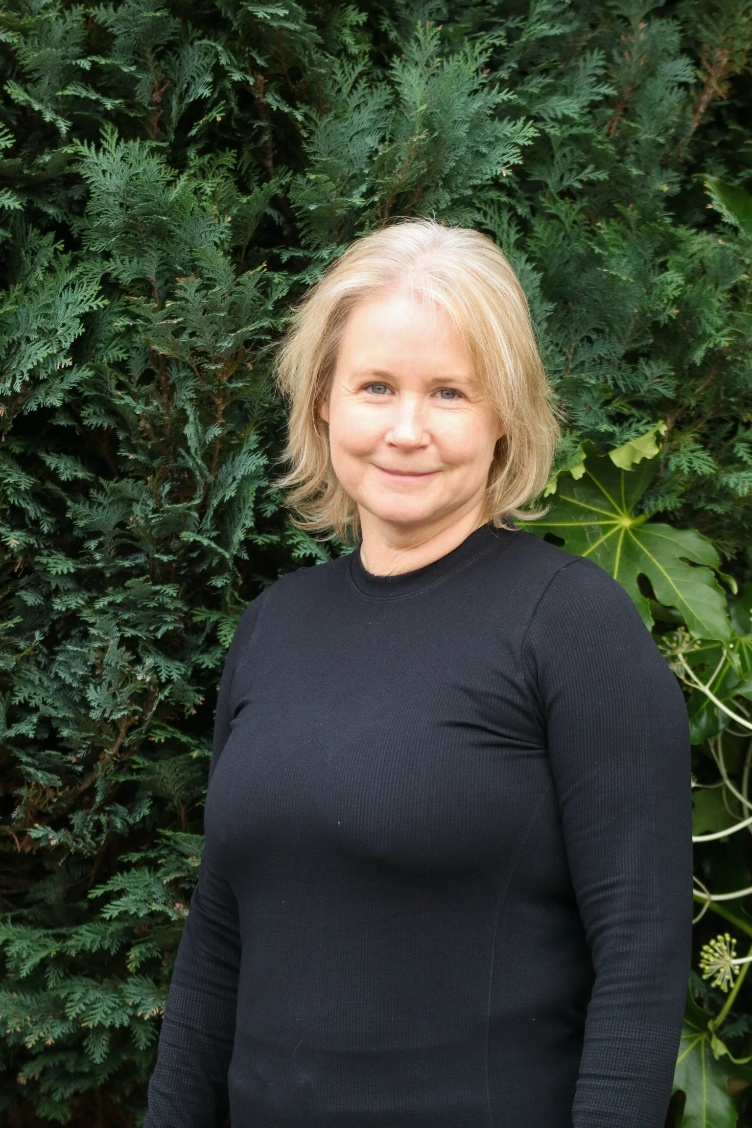 A woman with blonde hair wearing a black top standing in front of a green bush.
