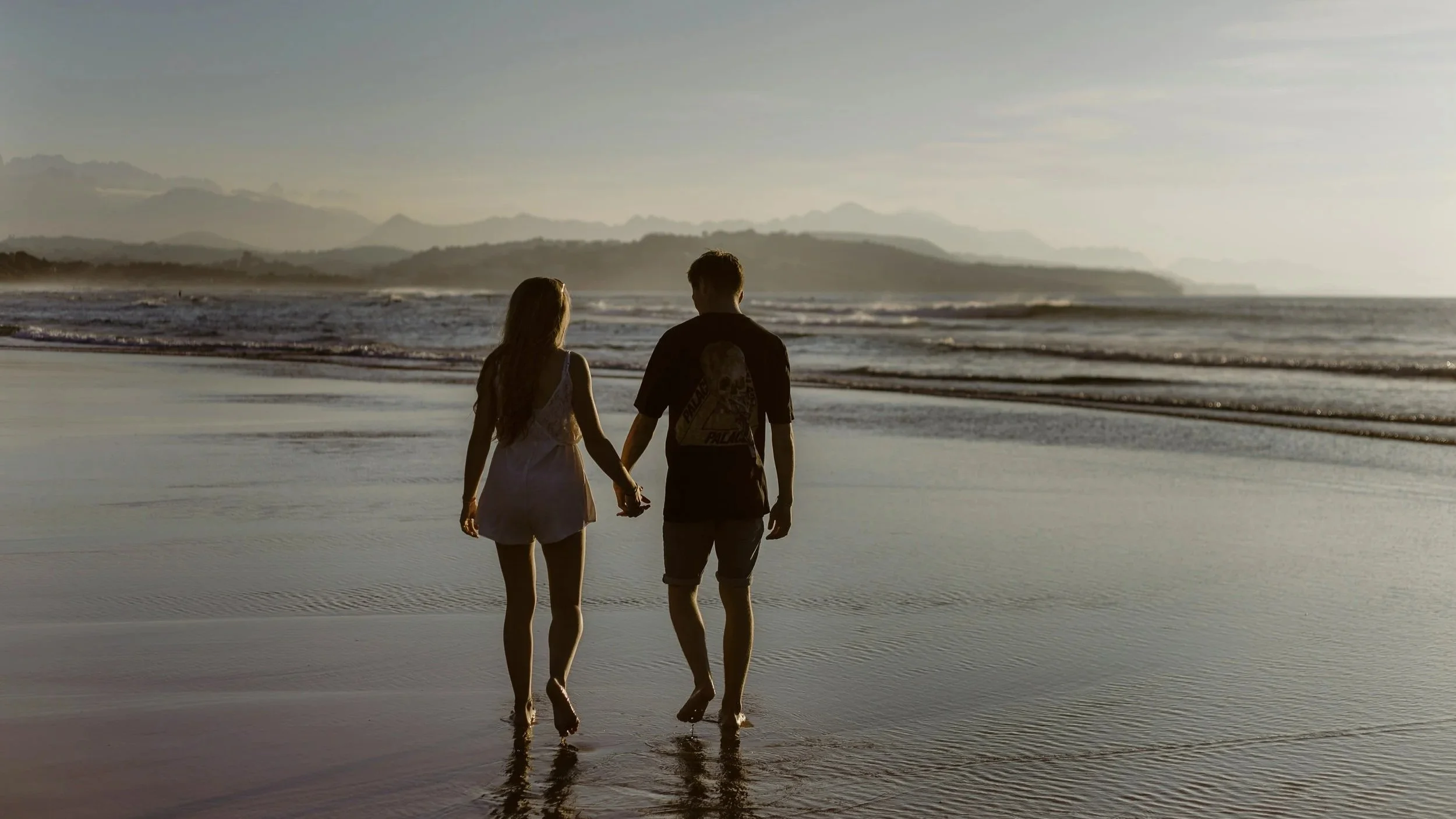A young couple holding hands walking along the shoreline at sunset on a beach, with mountains in the distance.