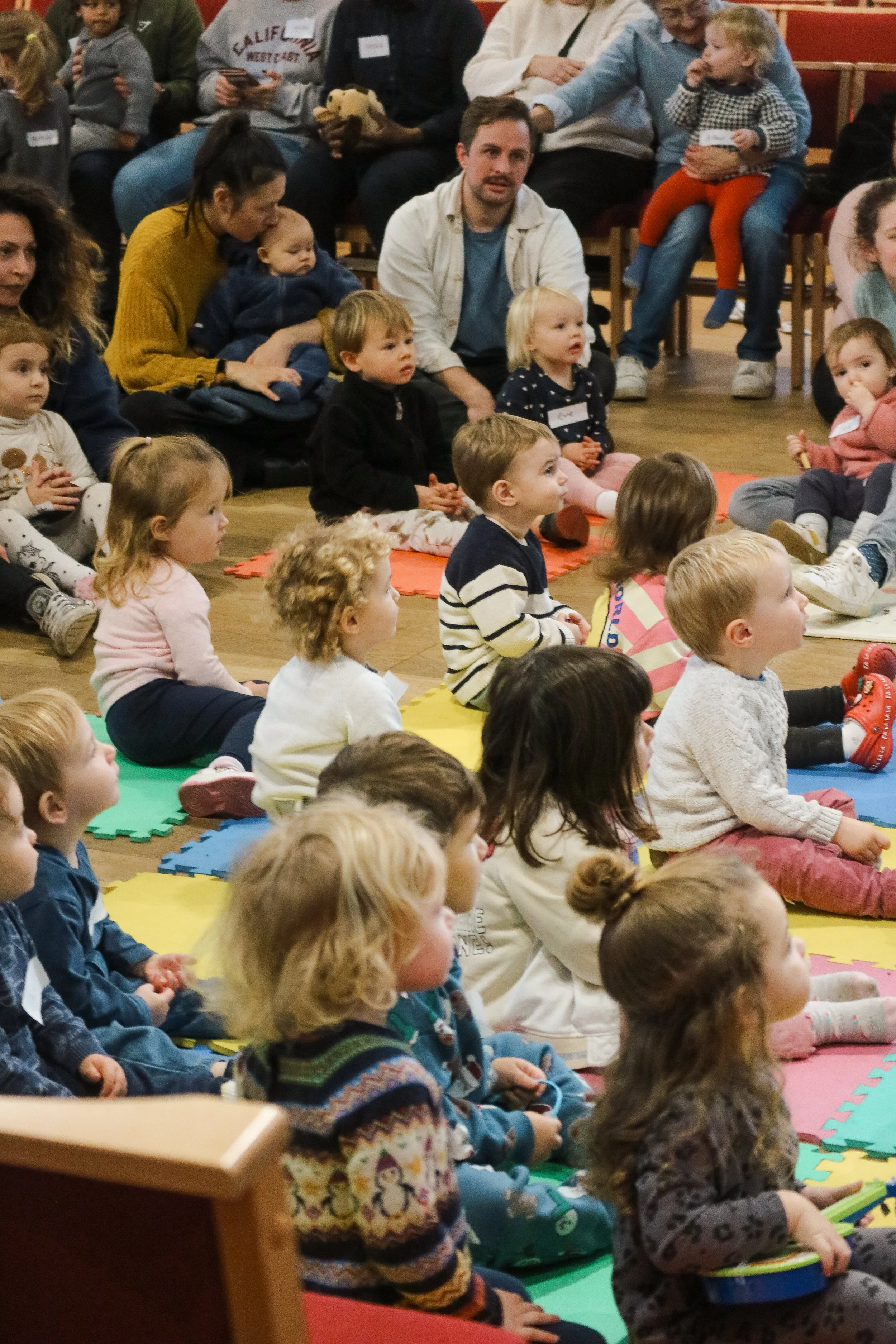 A group of young children sitting on colorful foam mats, facing forward, attending an indoor event. Several adults are seated behind them, some holding children or toys, in a room with wooden floors and red chairs.