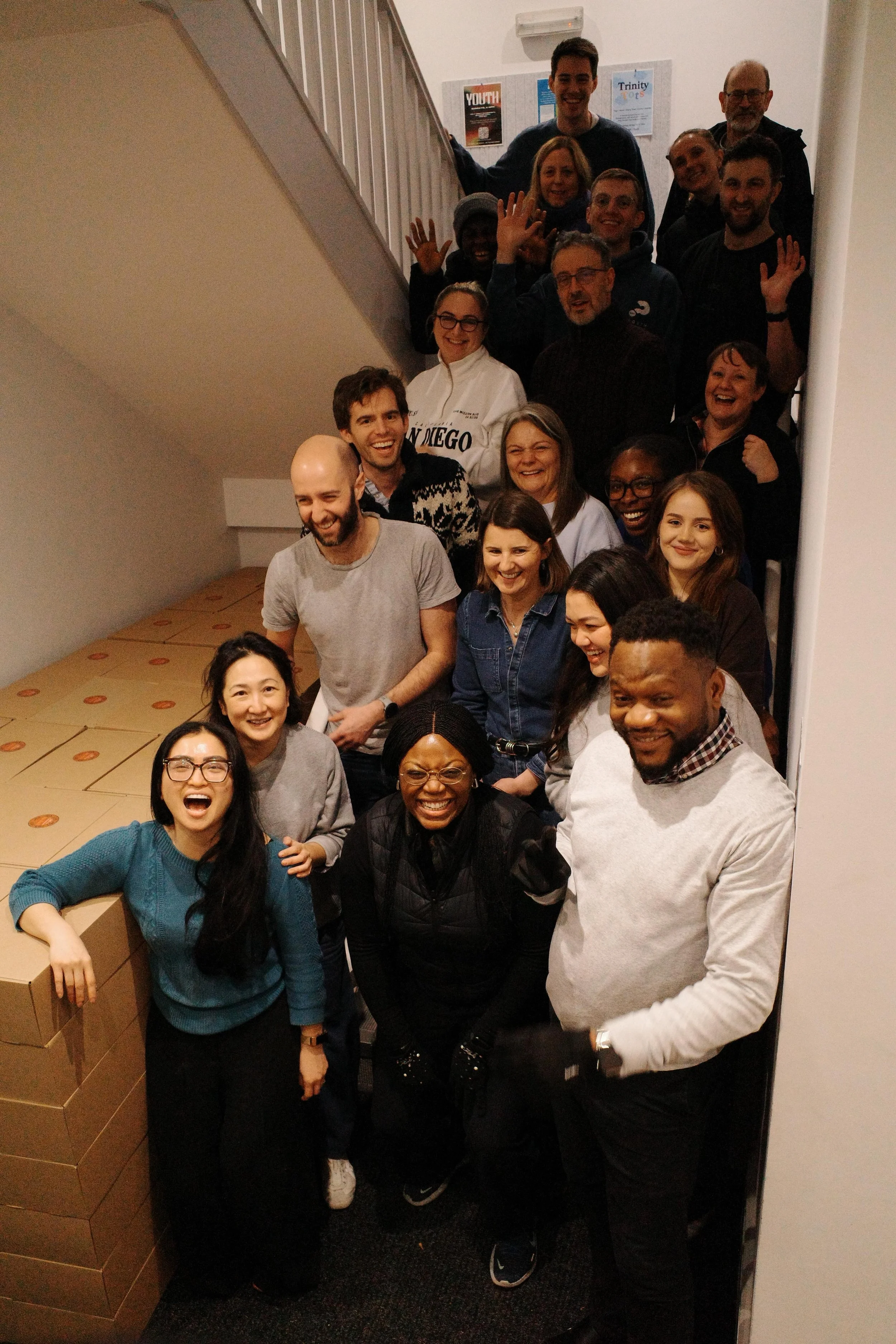 A group of people gathered on a staircase, smiling and waving at the camera, with some standing and others sitting around boxes in a room with white walls and posters.