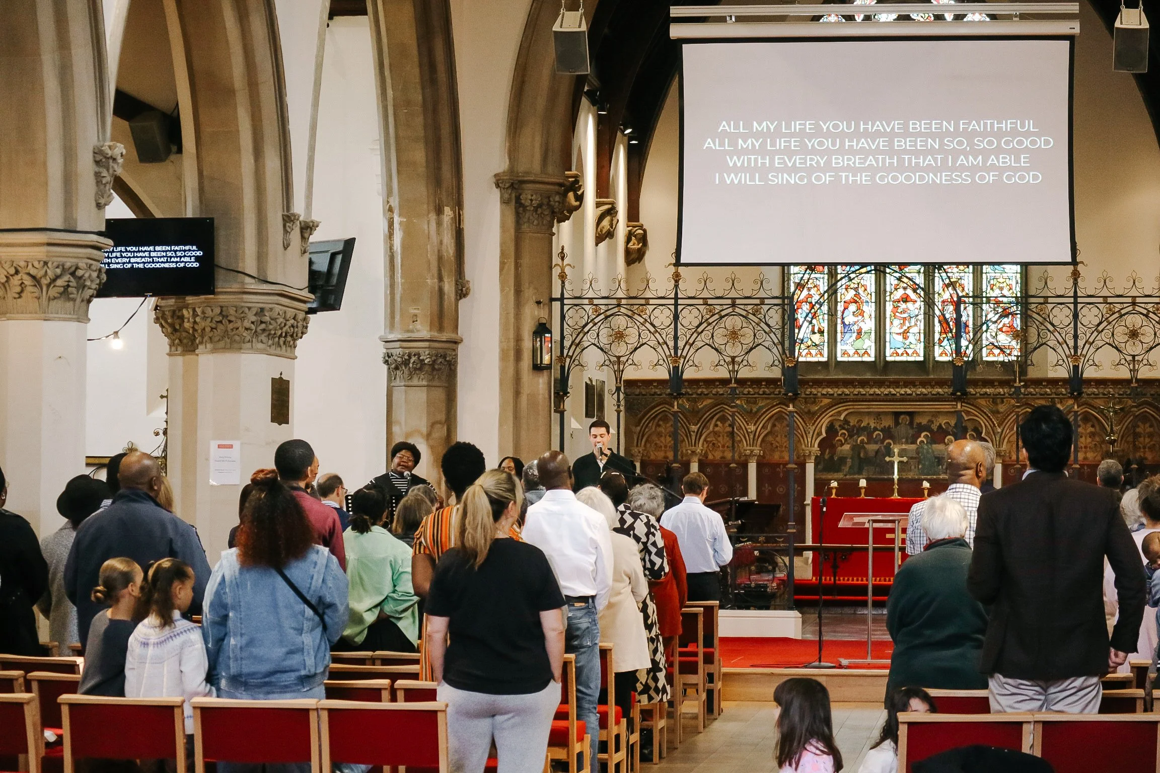 Inside a church with a congregation gathered for worship, a large screen displays lyrics of a hymn, and a man is speaking or singing at a microphone. The church has stained glass windows and ornate architectural details.