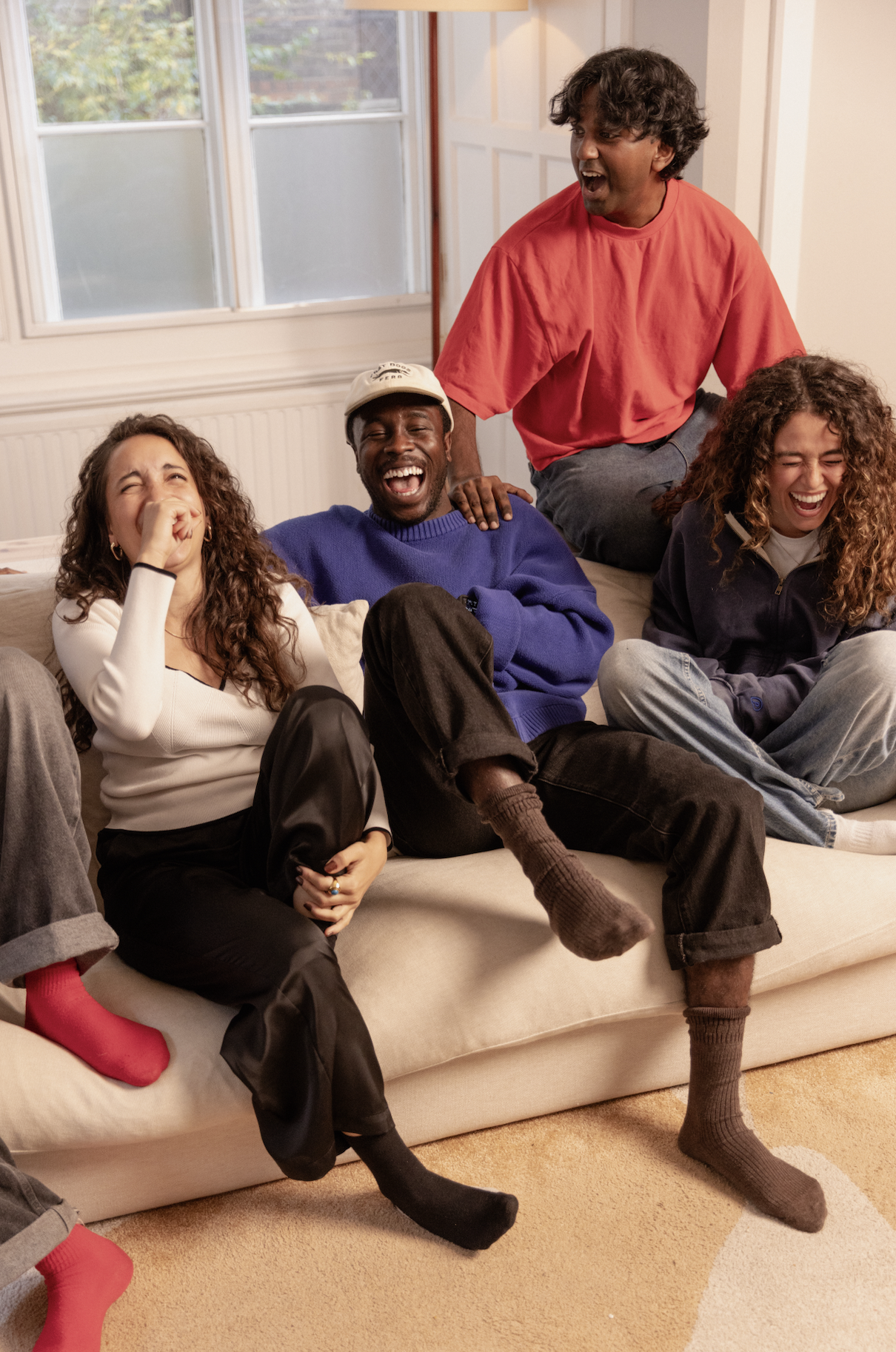 A group of five young people sitting on a beige sofa in a living room, laughing and enjoying each other's company, with large windows in the background.