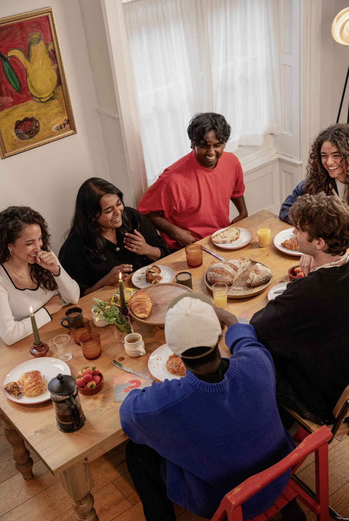 A group of seven people gathered around a wooden dining table, smiling and having breakfast with bread, croissants, strawberries, coffee, orange juice, and a cake. The setting is a bright room with white curtains, artwork on the wall, and a mix of casual and semi-formal attire.
