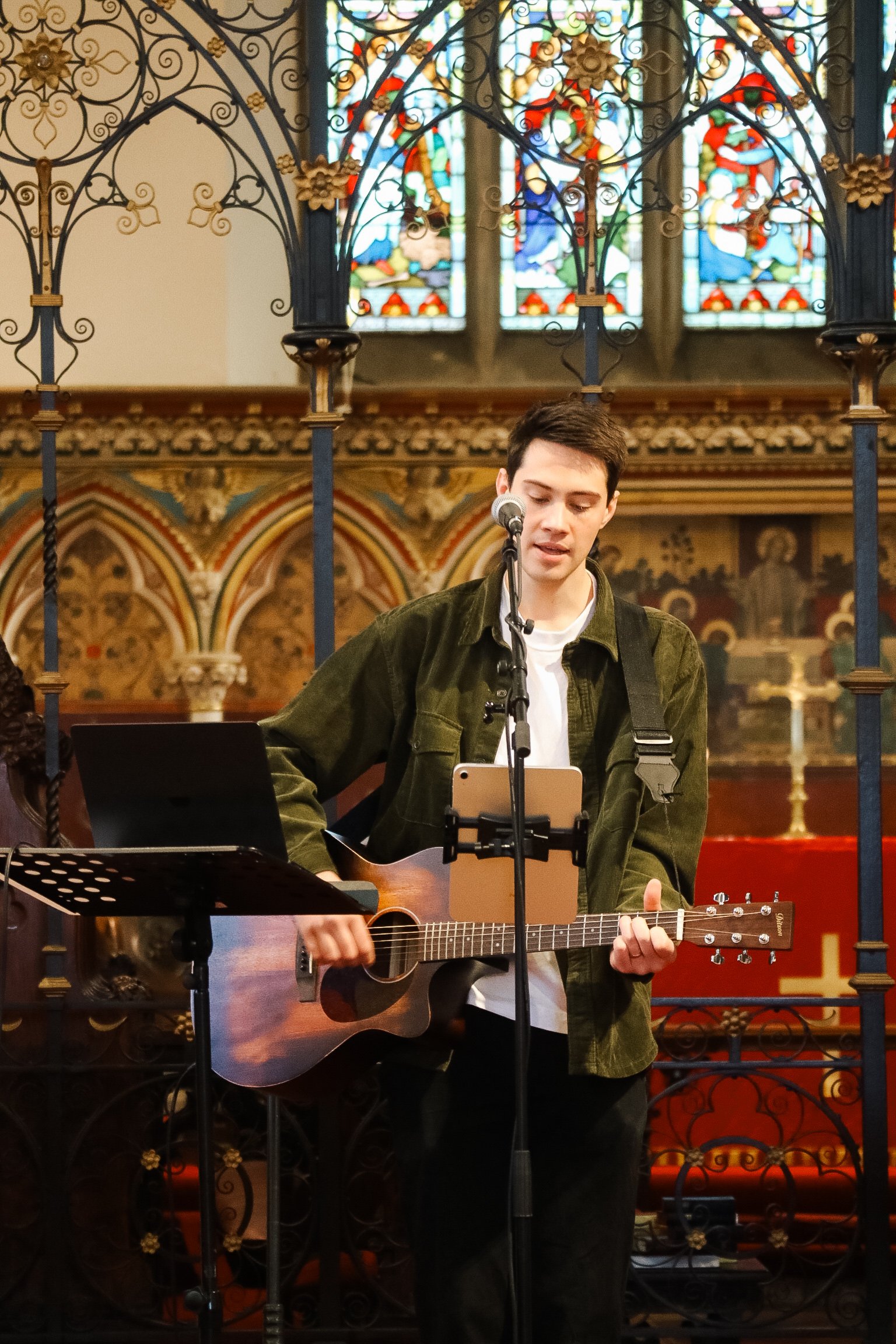 A young man with short dark hair, wearing a white shirt and green jacket, playing an acoustic guitar and singing into a microphone in a church with stained glass windows in the background.