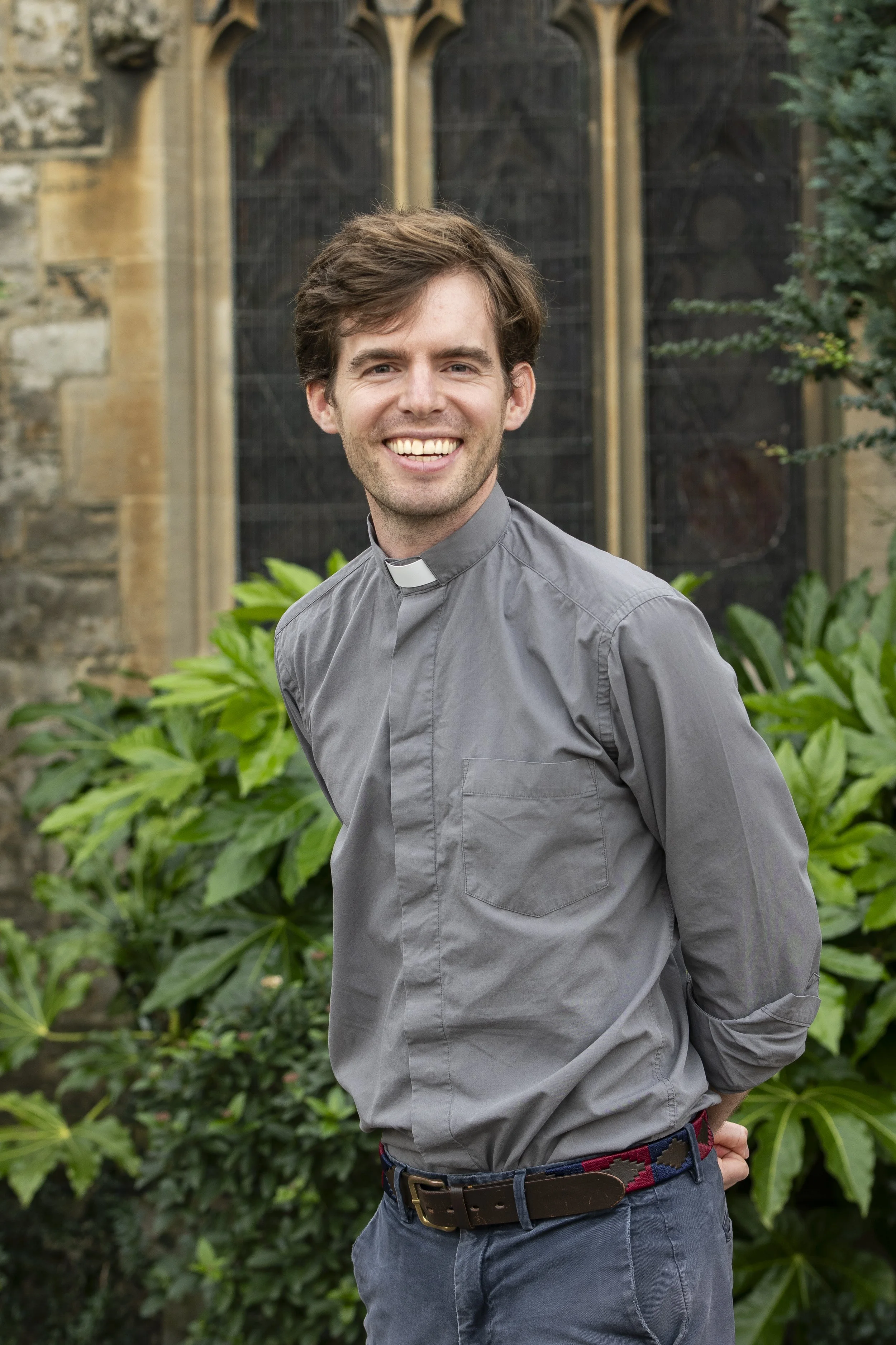 A smiling man dressed as a priest in a gray clerical shirt with a white collar standing outdoors amidst lush greenery and plants, with a historic stone building and a large Gothic window in the background.