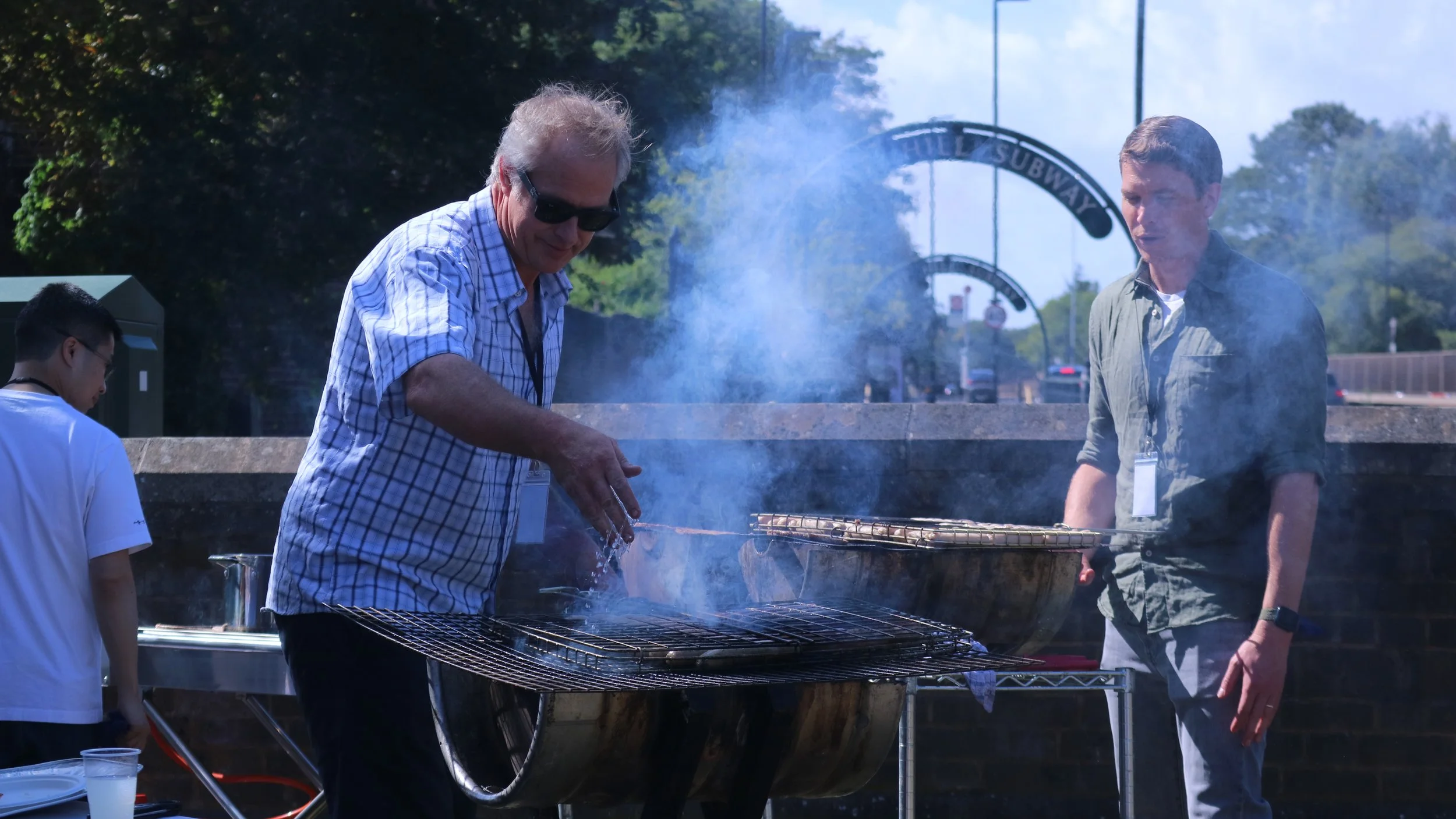 Three men and a boy grilling food on an outdoor barbecue grill, with puffing smoke and trees in the background.