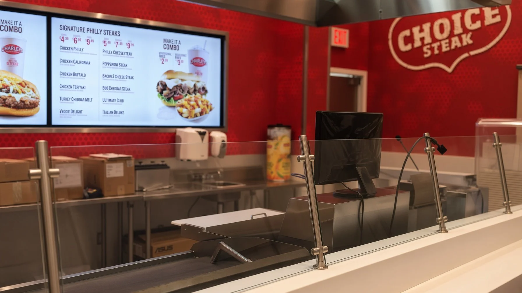 Counter area at a fast food restaurant with a digital menu screen displaying various combo meals and steak options, behind a glass shield, with a cash register and some boxes on the counter, and a red wall with a large sign that says 'Choice Steak'.