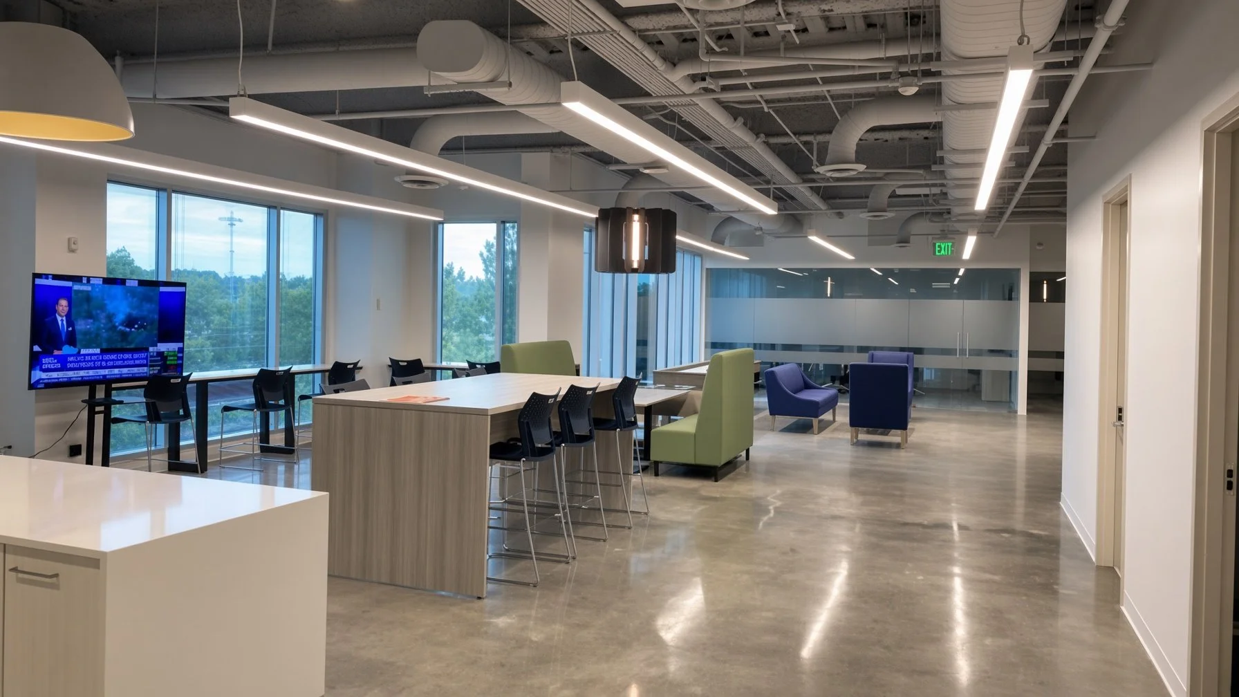 Open office lounge with a large window, TV, chairs, and lounge seating. Modern lighting, exposed ceiling ducts, and polished concrete floors.