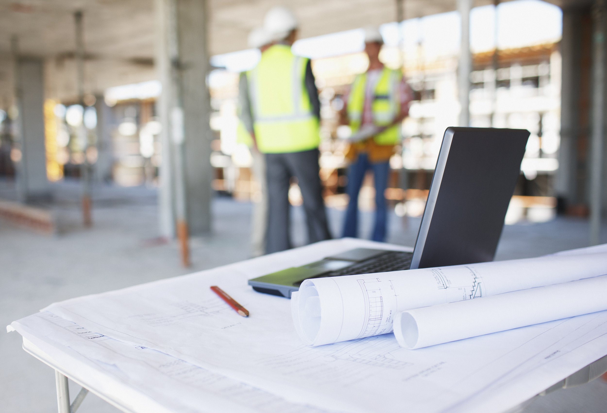 Architectural plans, a laptop, and a pencil on a white table in the foreground. In the background, two construction workers in safety vests and helmets are discussing at a construction site.
