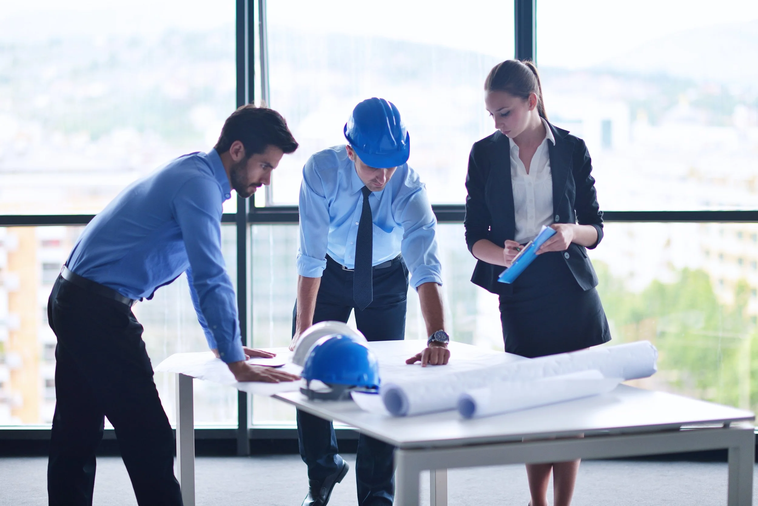 Three professionals, two men and one woman, examining blueprints and a rolled blueprint on a table in a bright office with large windows, with safety helmets on the table.