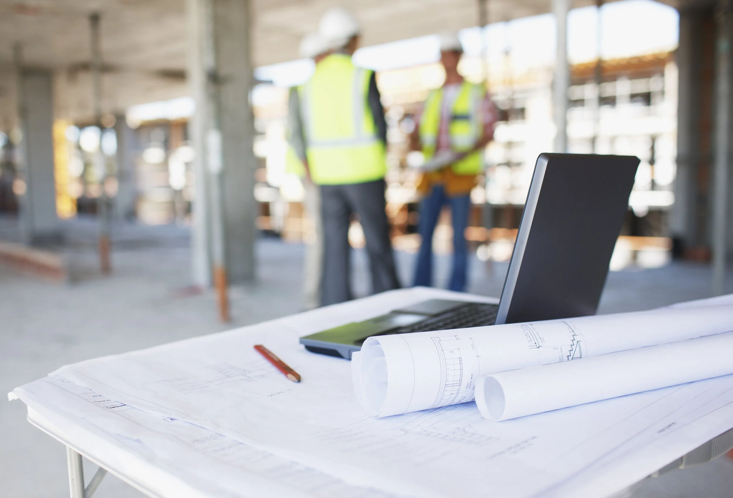 Architectural plans and blueprints on a white table with a laptop, in a construction site with two workers wearing safety vests and helmets in the background.