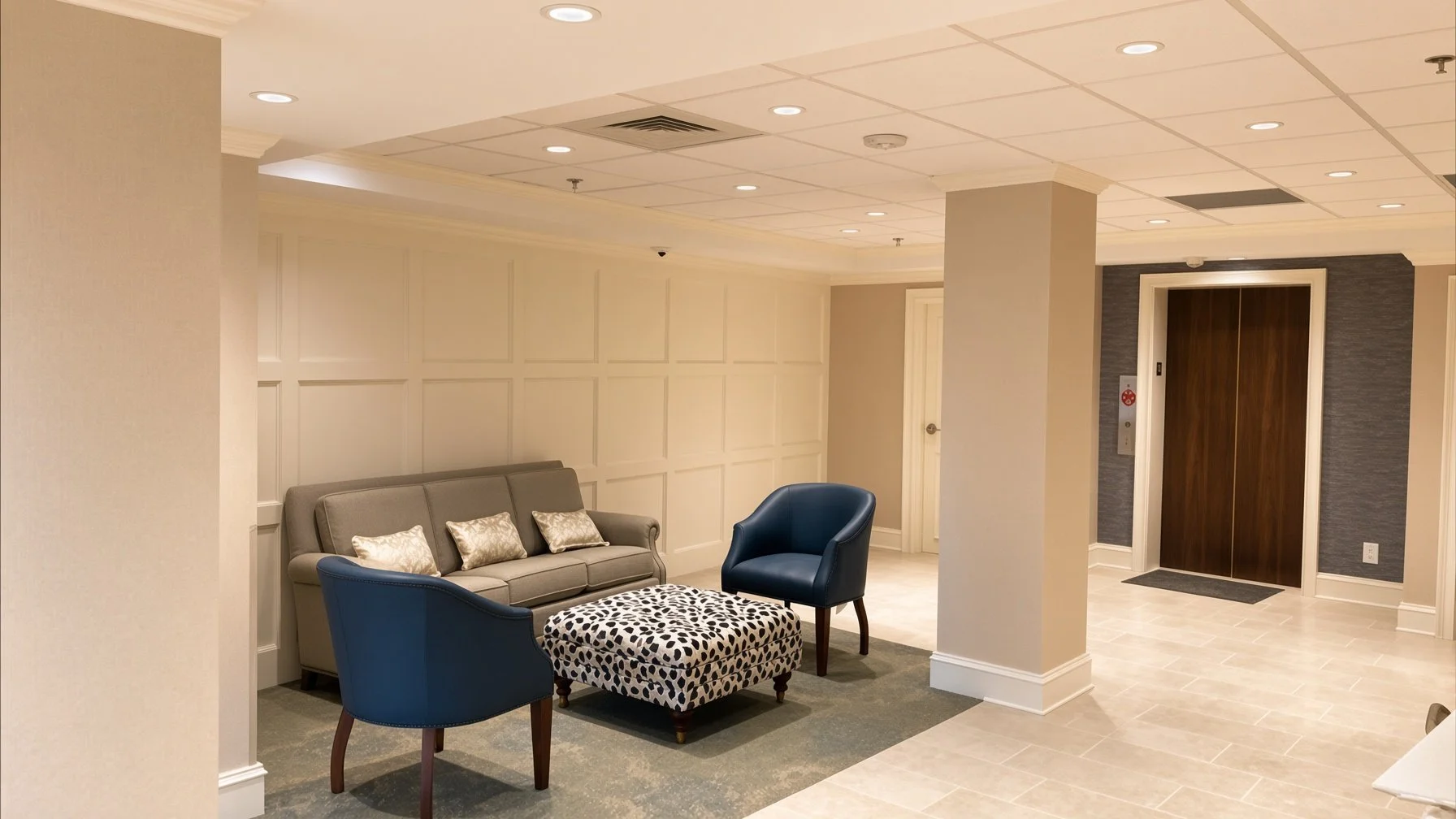 Hotel lobby area with seating including a gray sofa with three pillows, a blue armchair, and a cow-print ottoman, with cream-colored walls, a corner column, and an elevator with dark wood doors.