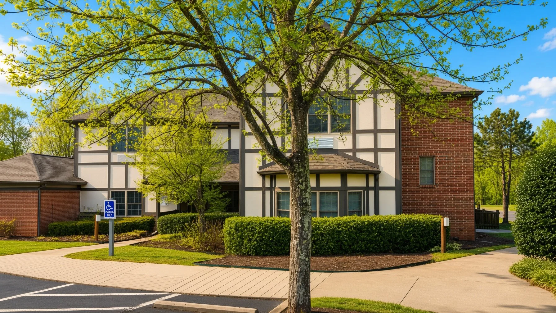 A three-story townhouse with white and black half-timbered siding, red brick walls, and multiple windows, surrounded by trees, bushes, and a paved walkway, with a handicapped parking sign in the foreground.