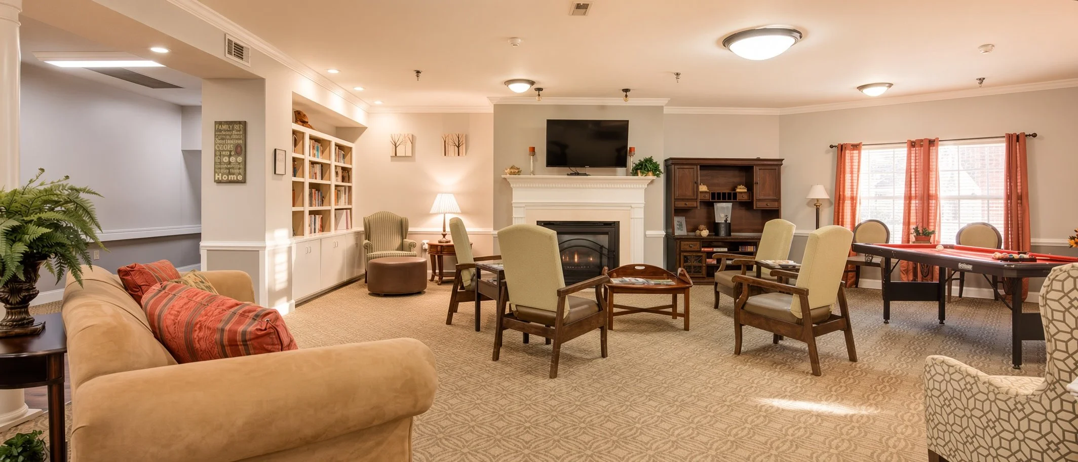 Comfortable living room with beige sofa, armchairs, fireplace, TV mounted above fireplace, bookshelf, table with lamp, window with orange curtains, pool table, plant, and various decorative items.