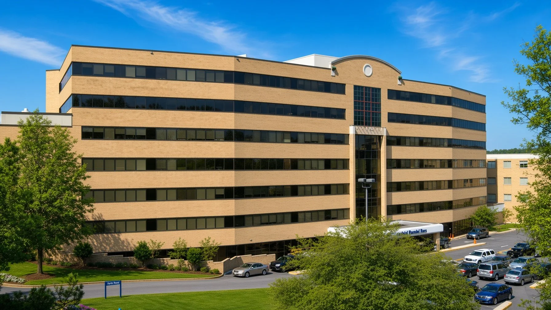 View of a large multi-story office building with beige brick exterior, black tinted windows, and a decorative dome on top, surrounded by trees and a parking lot.