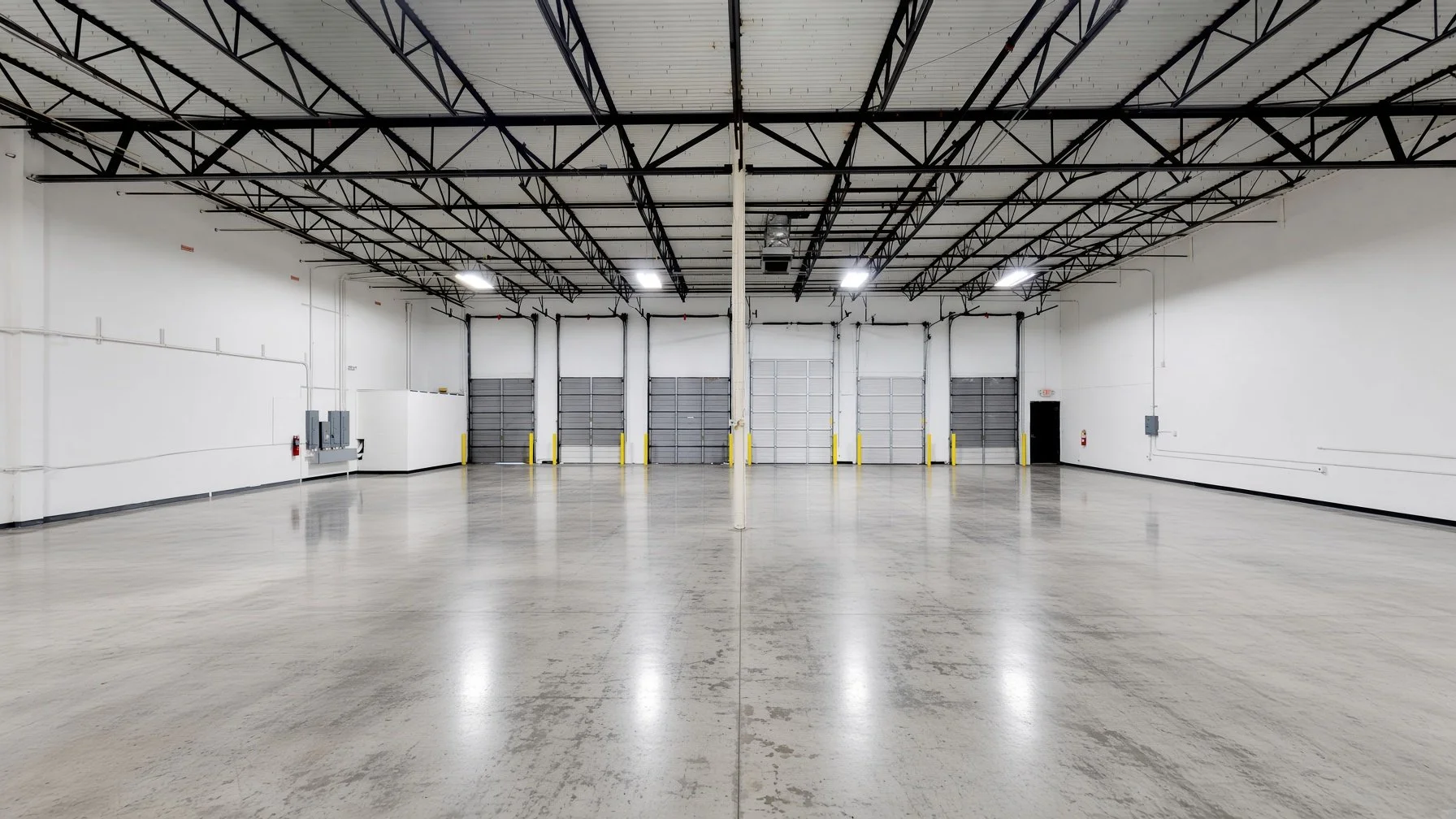 Empty warehouse or industrial space with a polished concrete floor, white walls, and a high ceiling with black metal beams. There are multiple garage-style doors at the back, some with yellow safety bumpers, and bright overhead lighting.