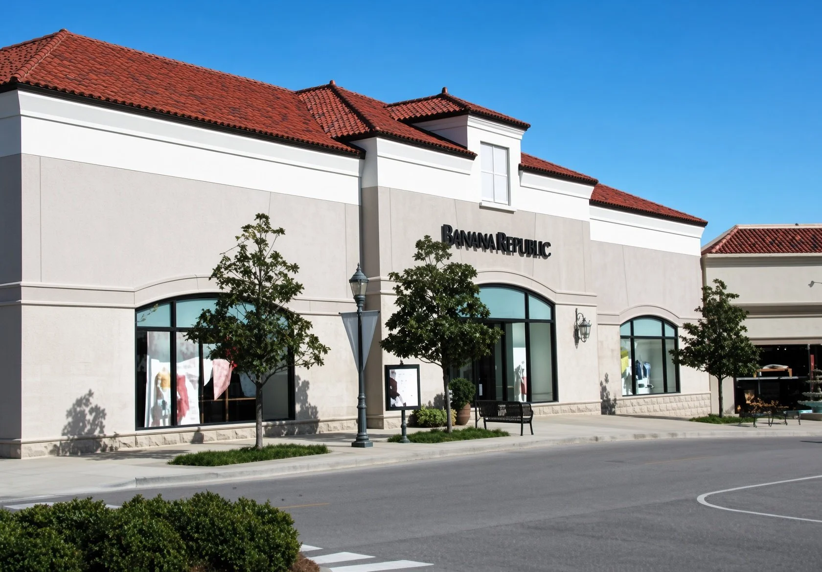 Exterior of a shopping mall with a Banana Republic store, red-tiled roof, large windows, trees, benches, and street lamps, under a clear blue sky.