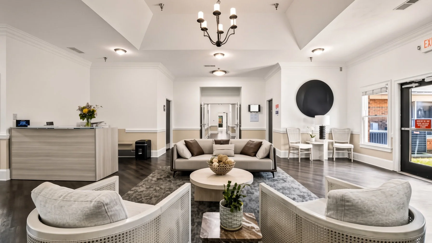 Lobby area with cream-colored walls, dark wood flooring, and modern lighting. Features include a beige sofa with brown and beige pillows, a round white coffee table with a basket of decorative balls, and white accent chairs. A reception desk with a flower arrangement and a small table with a lamp and plant are near the windows, which have white blinds and a red sign outside.