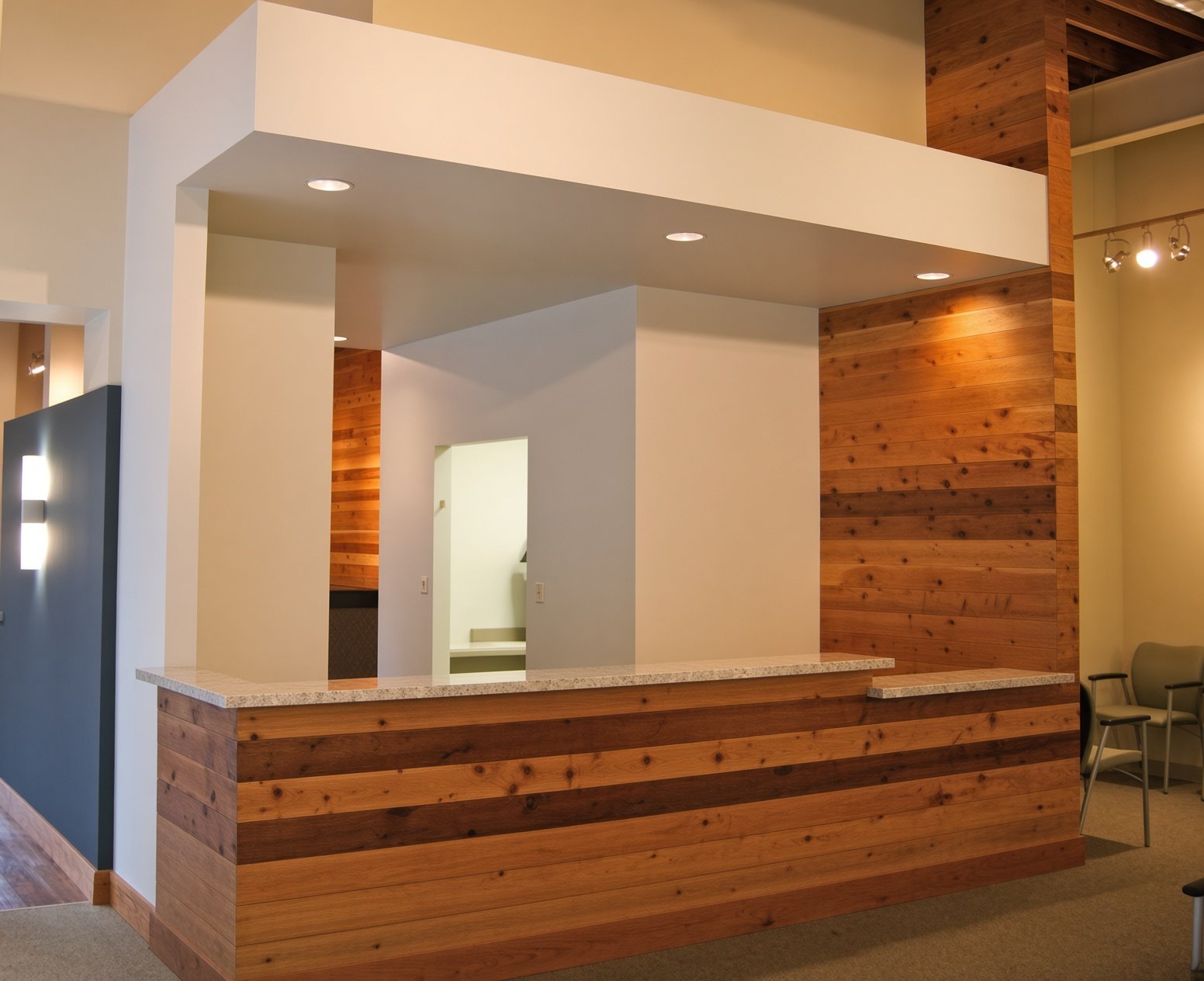Interior of a reception or lobby area with a wood-paneled counter and wall, white walls, recessed ceiling lights, and a small seating area with chairs.
