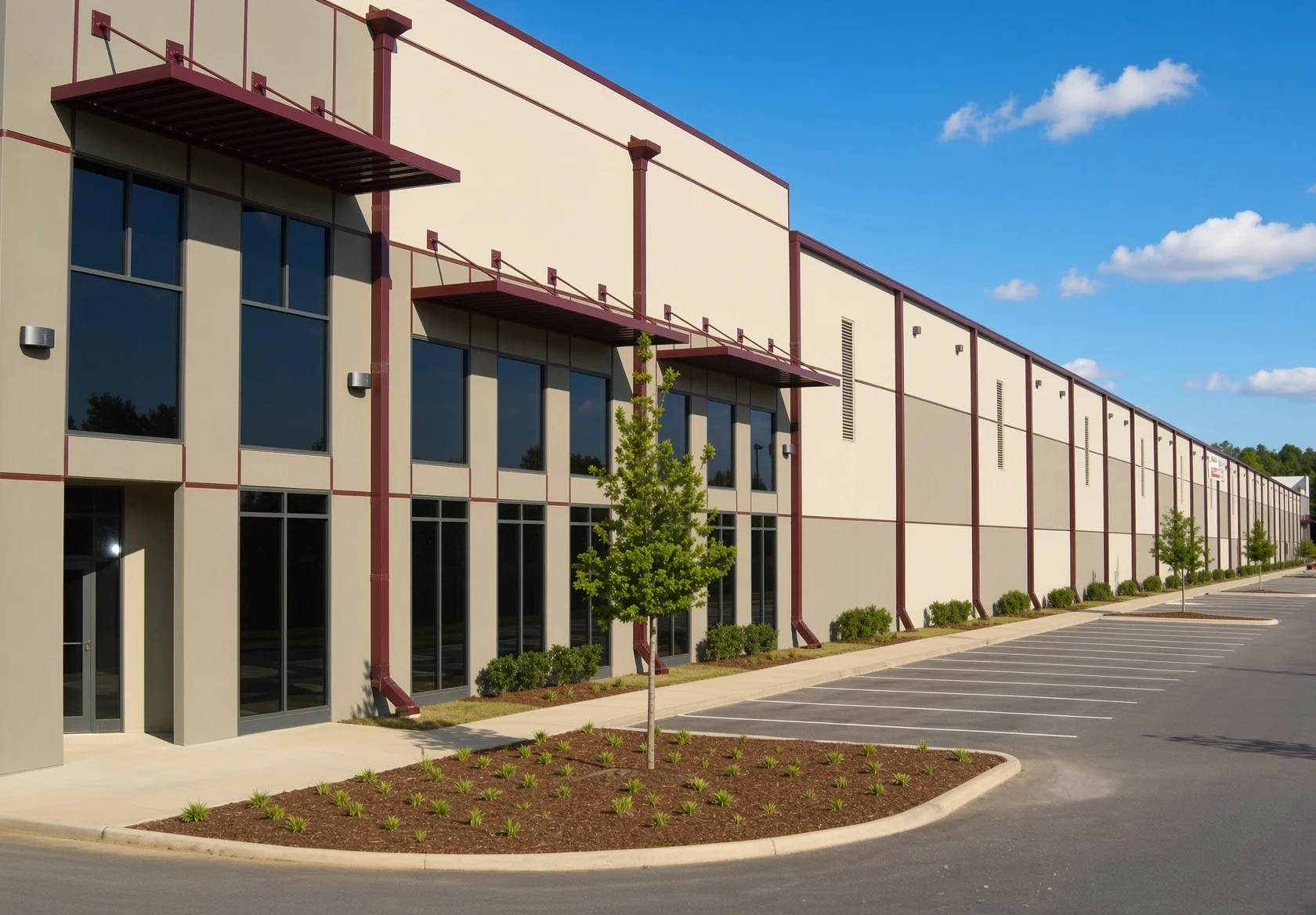 A large, modern commercial building with beige walls, red accents, large windows, and a parking lot with marked spaces. Young trees and small bushes line the sidewalk in front.