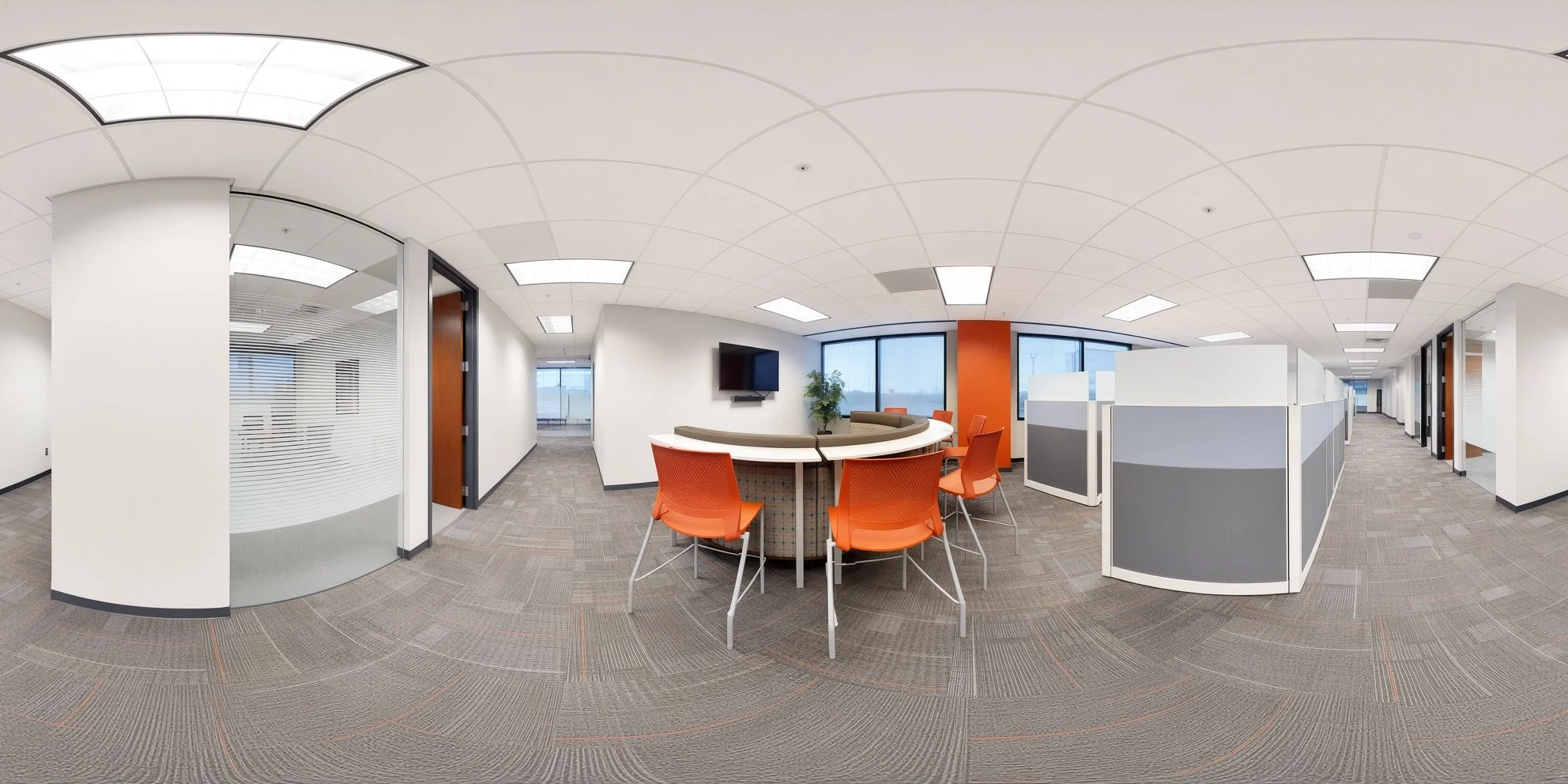 Office lounge area with round table, orange chairs, a wall-mounted TV, large windows, and cubicles.