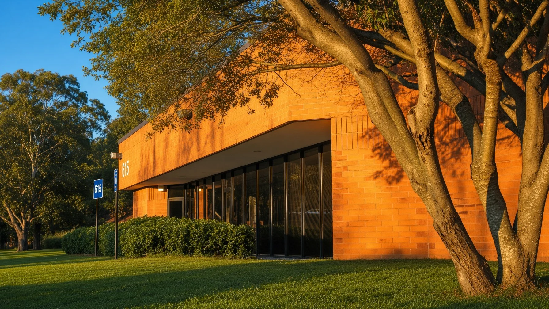 A brick building with large glass windows and the number 615 on the wall, surrounded by green grass and trees in the sunlight.