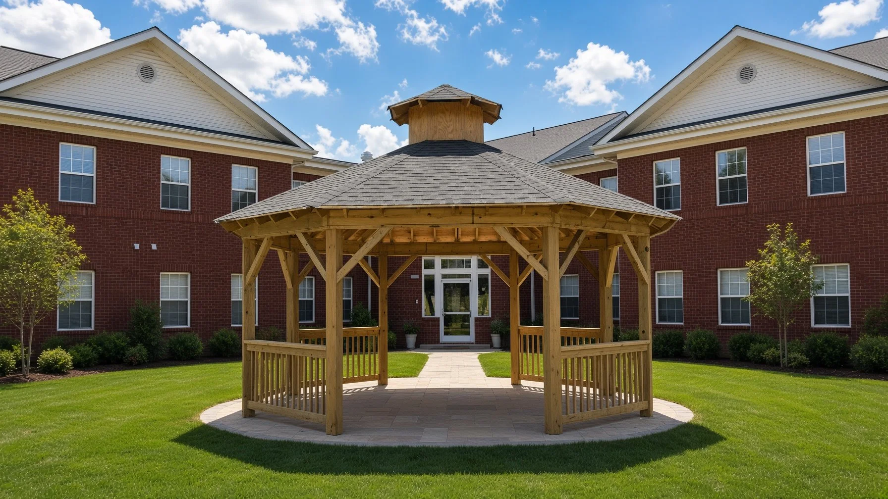 A wooden gazebo with a shingled roof in a green grassy backyard, with a brick apartment building in the background, under a blue sky with scattered clouds.