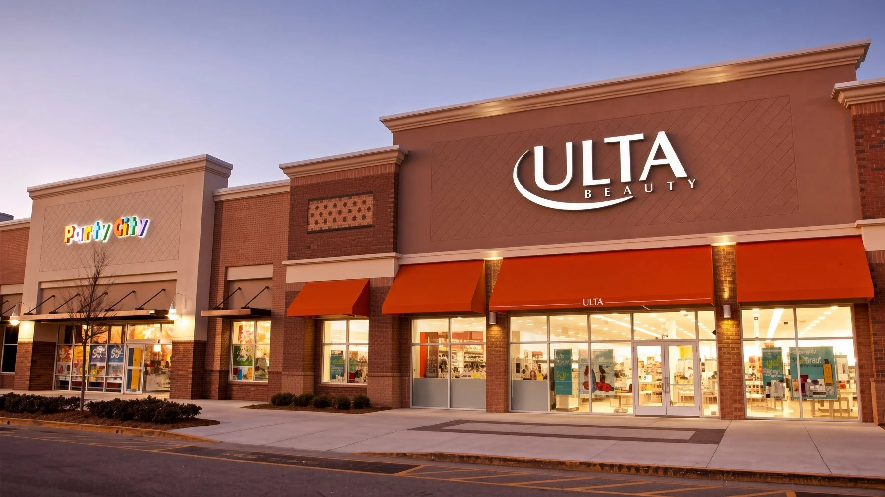 Shopping plaza storefront featuring ULTA Beauty and Party City stores with large glass windows and orange awnings, illuminated during dusk.