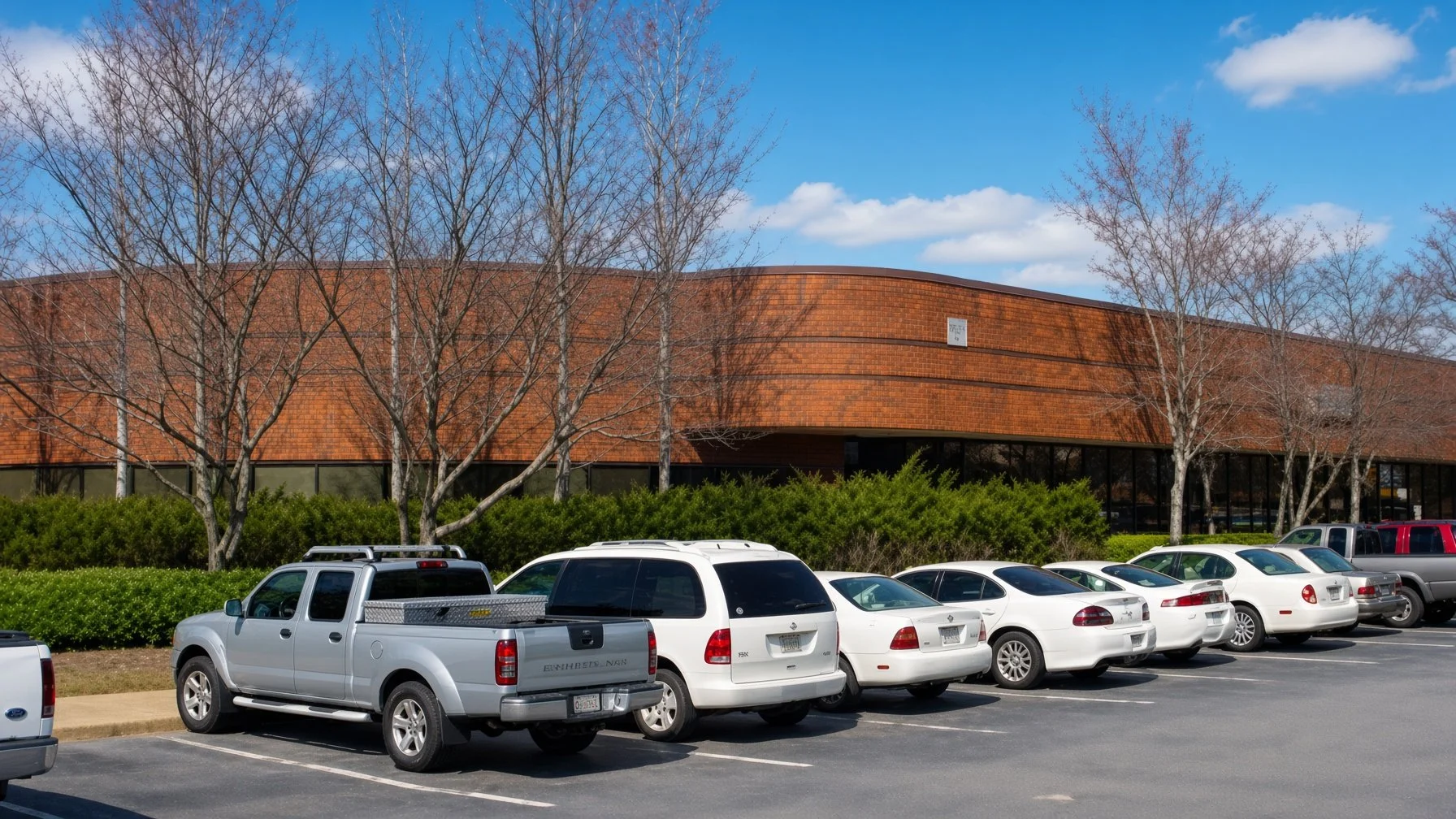 A parking lot with several white and silver cars parked in front of a large, curved brick building with leafless trees and green bushes surrounding it. The sky is blue with a few clouds.