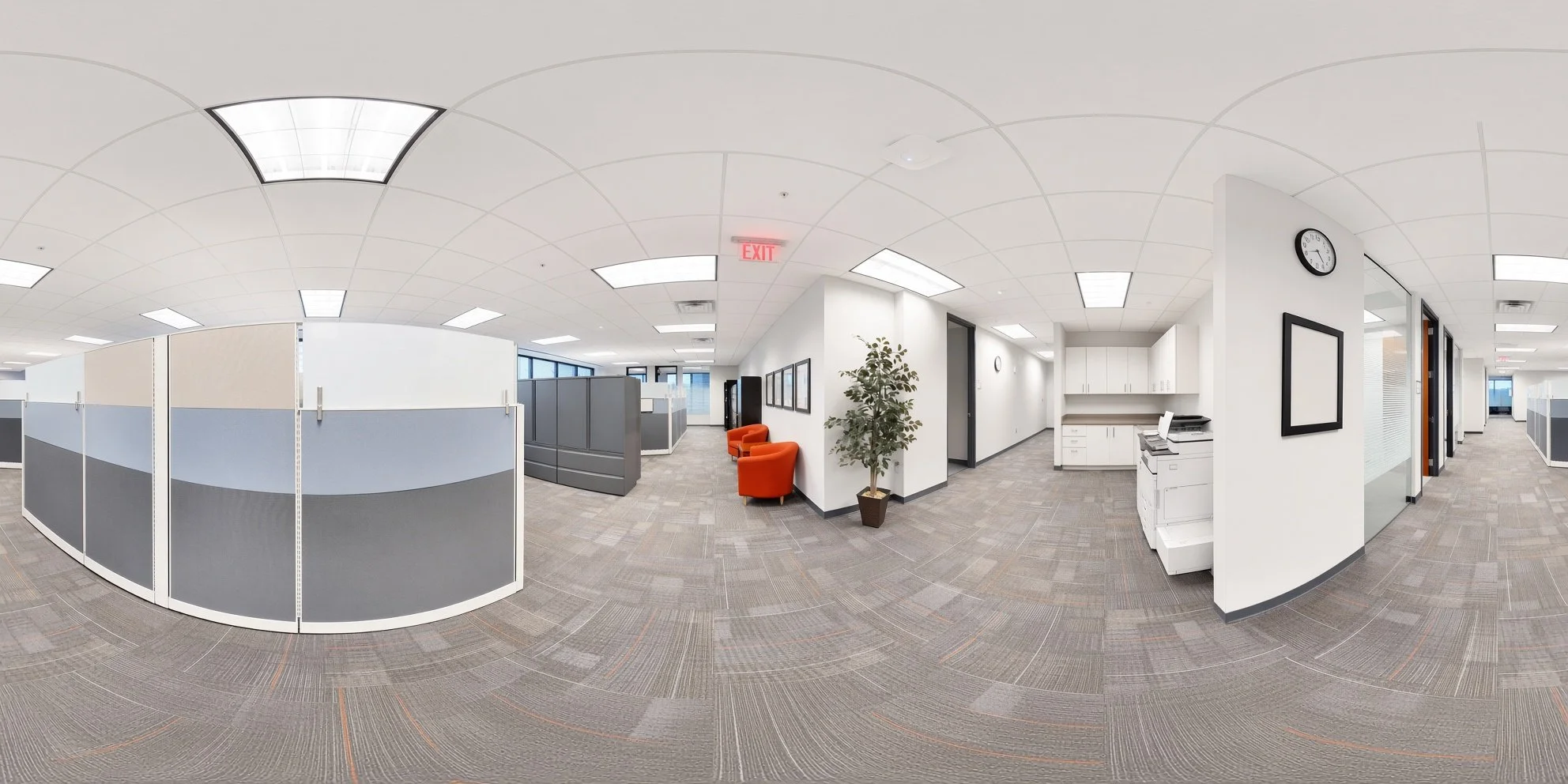 A modern, empty office space with cubicles, a small waiting area with orange chairs, and a kitchenette, illuminated by overhead fluorescent lights.