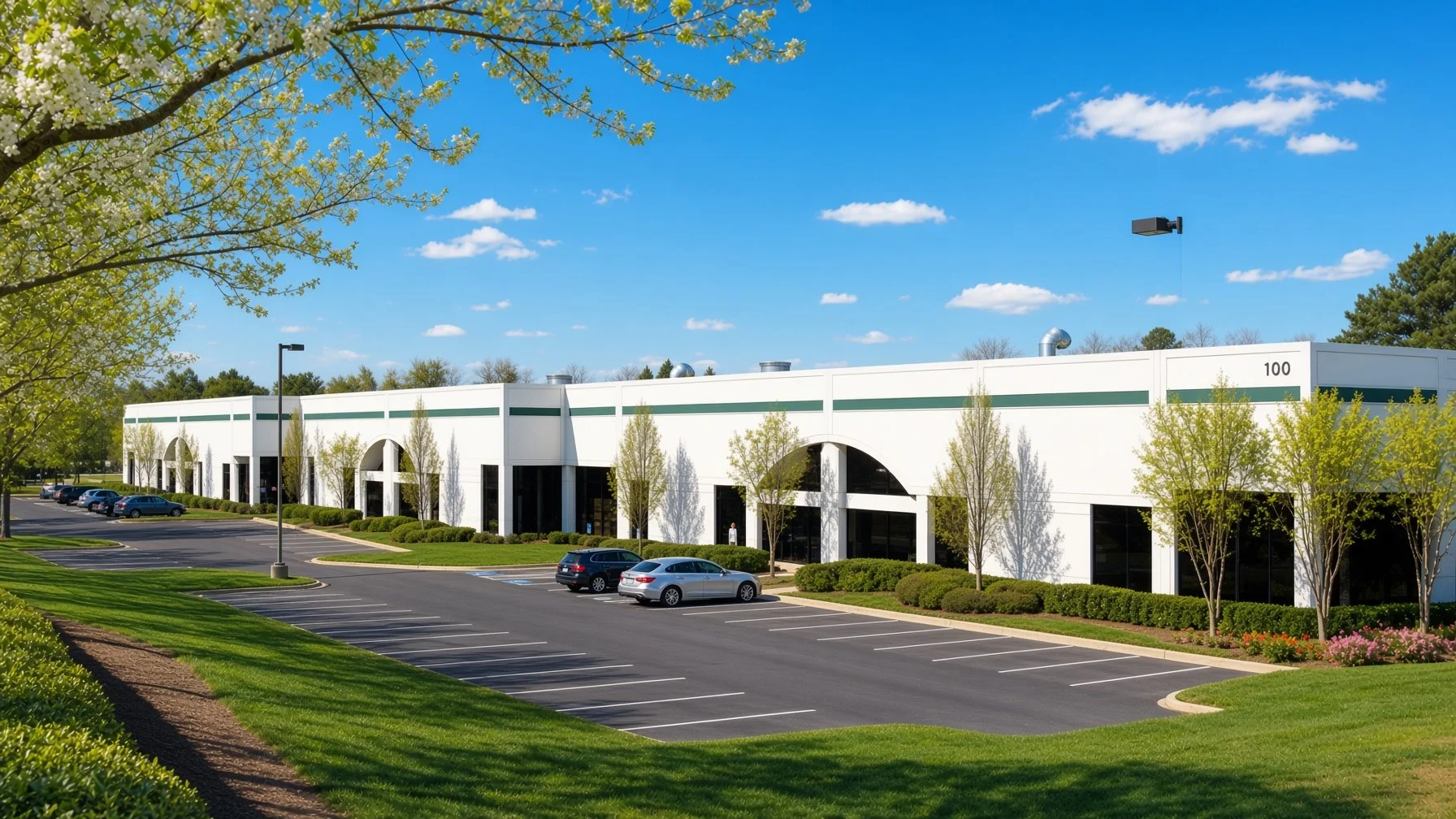 A commercial building with large glass windows and arched entrances, surrounded by a parking lot with several parked cars, green trees, shrubs, and blooming flowers on a bright, sunny day with a blue sky and scattered clouds.