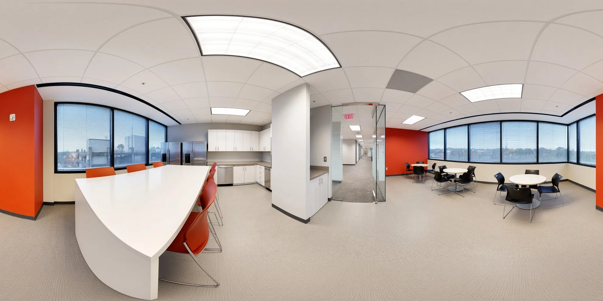 Interior of an office break room with large windows, white walls, red accent wall, and round tables with black chairs. Kitchen area with white cabinets, a refrigerator, and a microwave. Ceiling has square lights and a curved design.