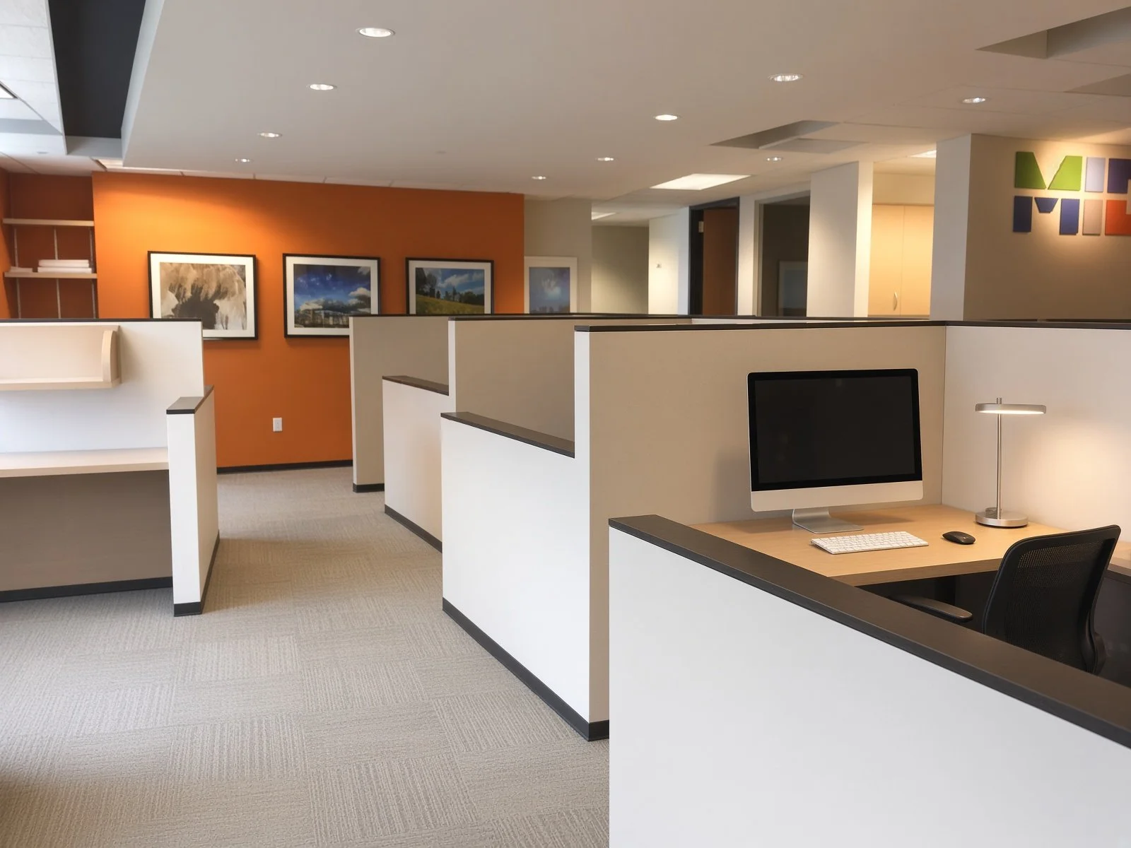 Empty office cubicles with a computer, desk lamp, and chairs, in a modern office space with artwork on the orange wall.