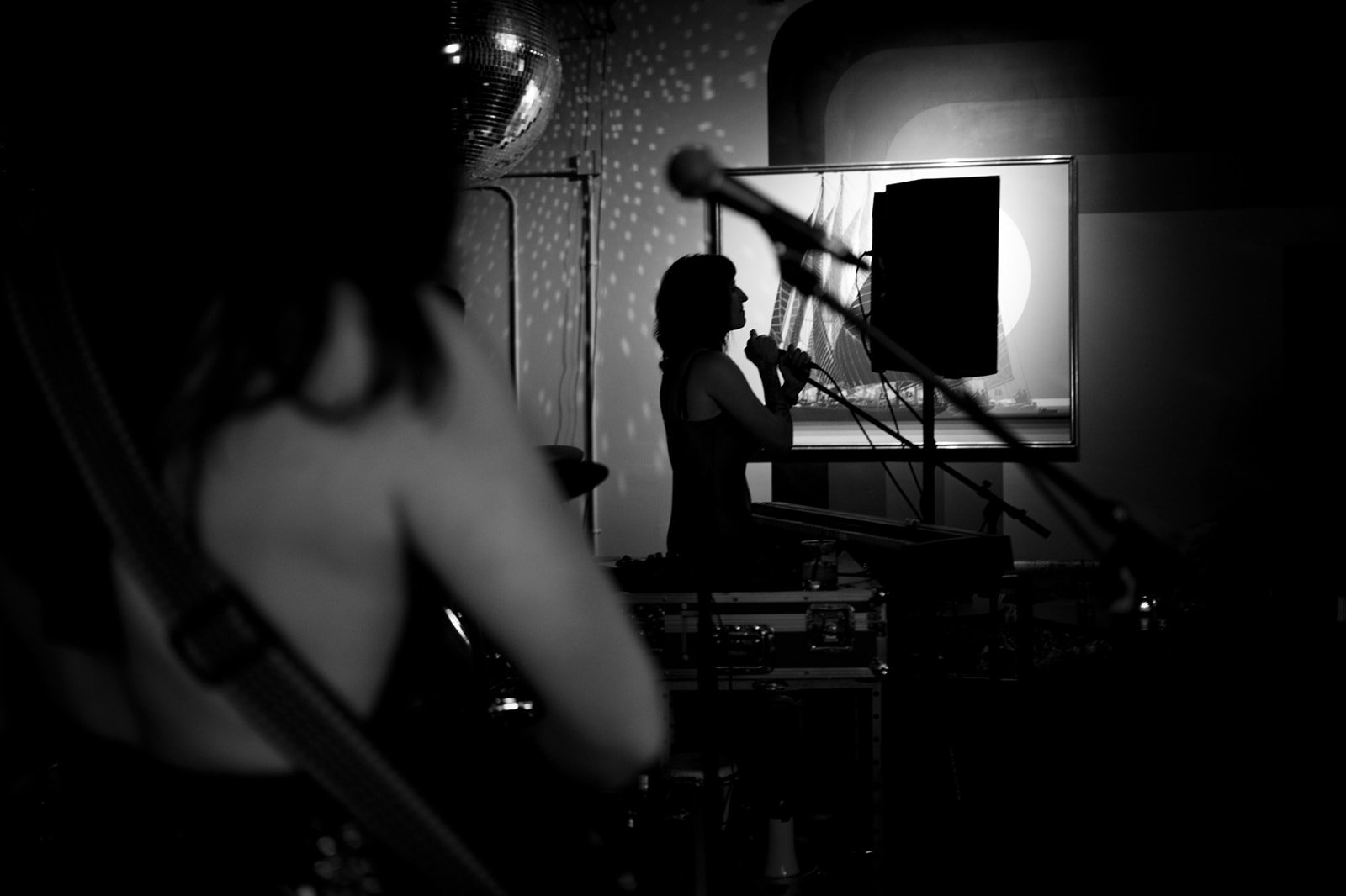 Black and white photo of a woman singing into a microphone during a performance, with a shadowed figure of a musician playing a guitar in the foreground and stage equipment visible.