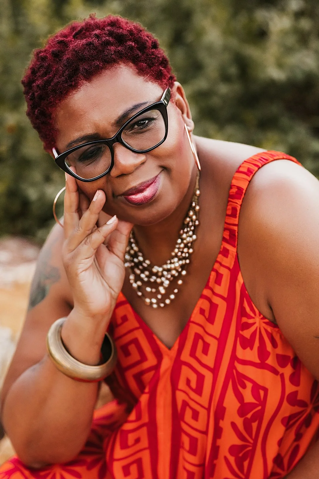 A woman with short curly red hair wearing black glasses, hoop earrings, a pearl necklace, a white bangle, and a red patterned dress is resting her face on her hand outdoors.