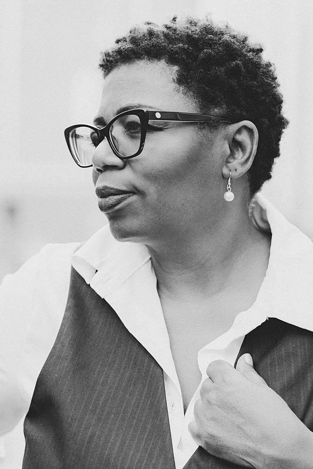 Side view of a woman with short curly hair wearing glasses and a pearl earring, dressed in a white shirt and dark vest. Black and white photograph.