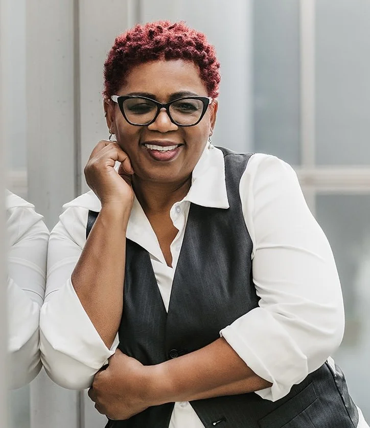 A woman with short, curly red hair and glasses, smiling and resting her chin on her hand, wearing a white shirt and dark vest, standing outdoors.