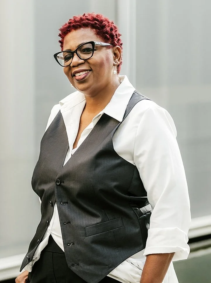A woman with short, curly red hair and black glasses smiling, wearing a white shirt with the sleeves rolled up and a black vest, standing indoors.