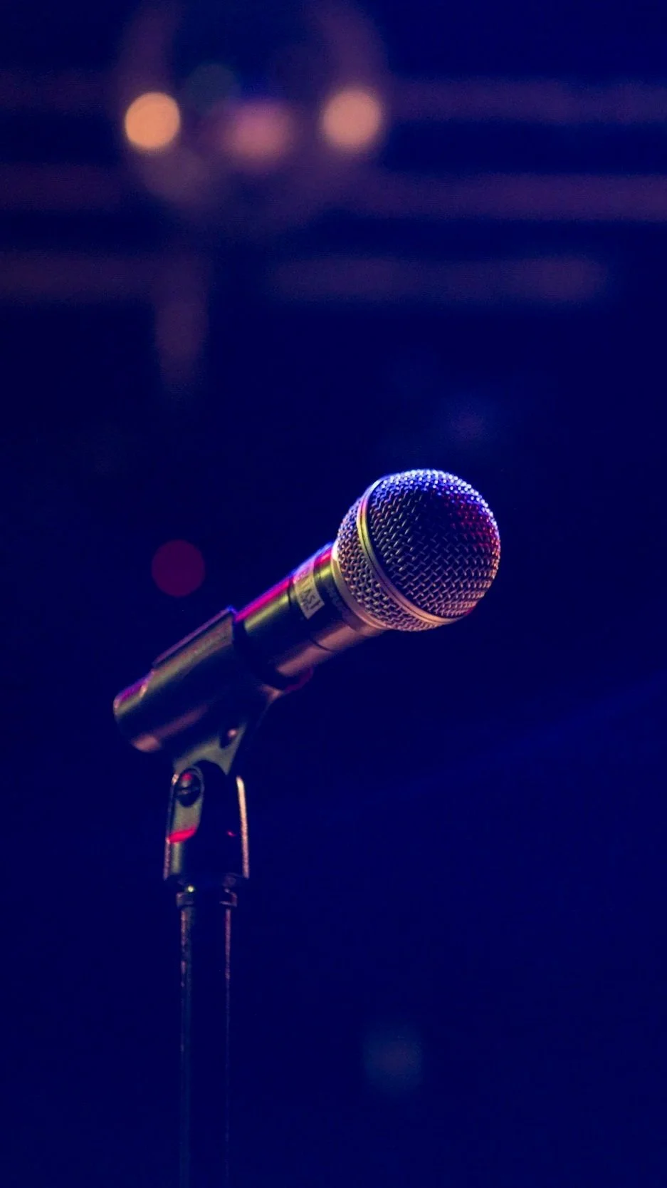 Close-up of a microphone on a stand, set against a dark background with blurred lights.
