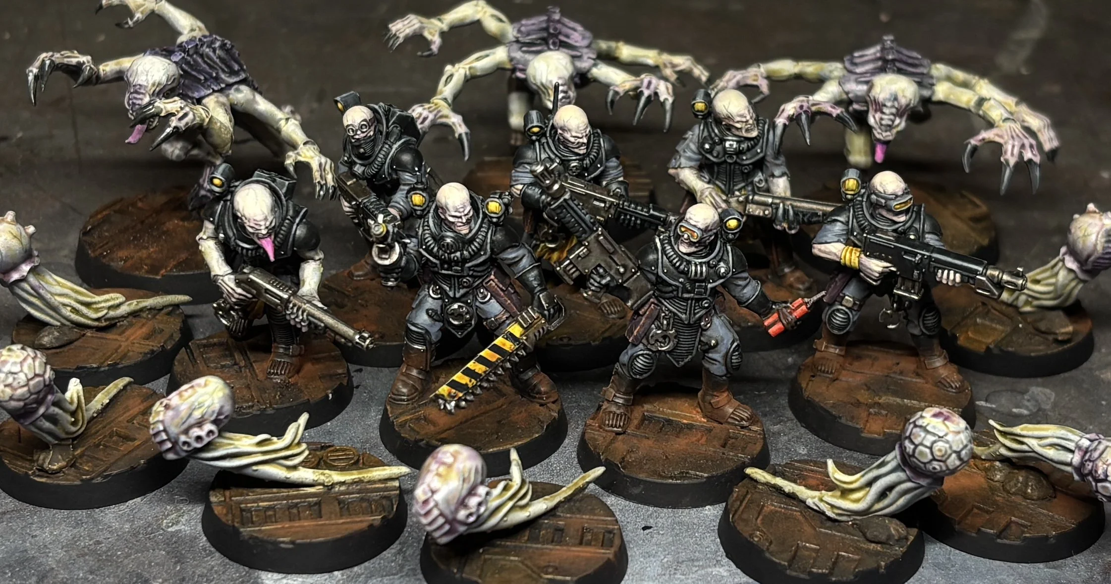 Miniature figurines of soldiers in black armor wielding guns, surrounded by alien creatures with elongated limbs and tails, on a textured base.