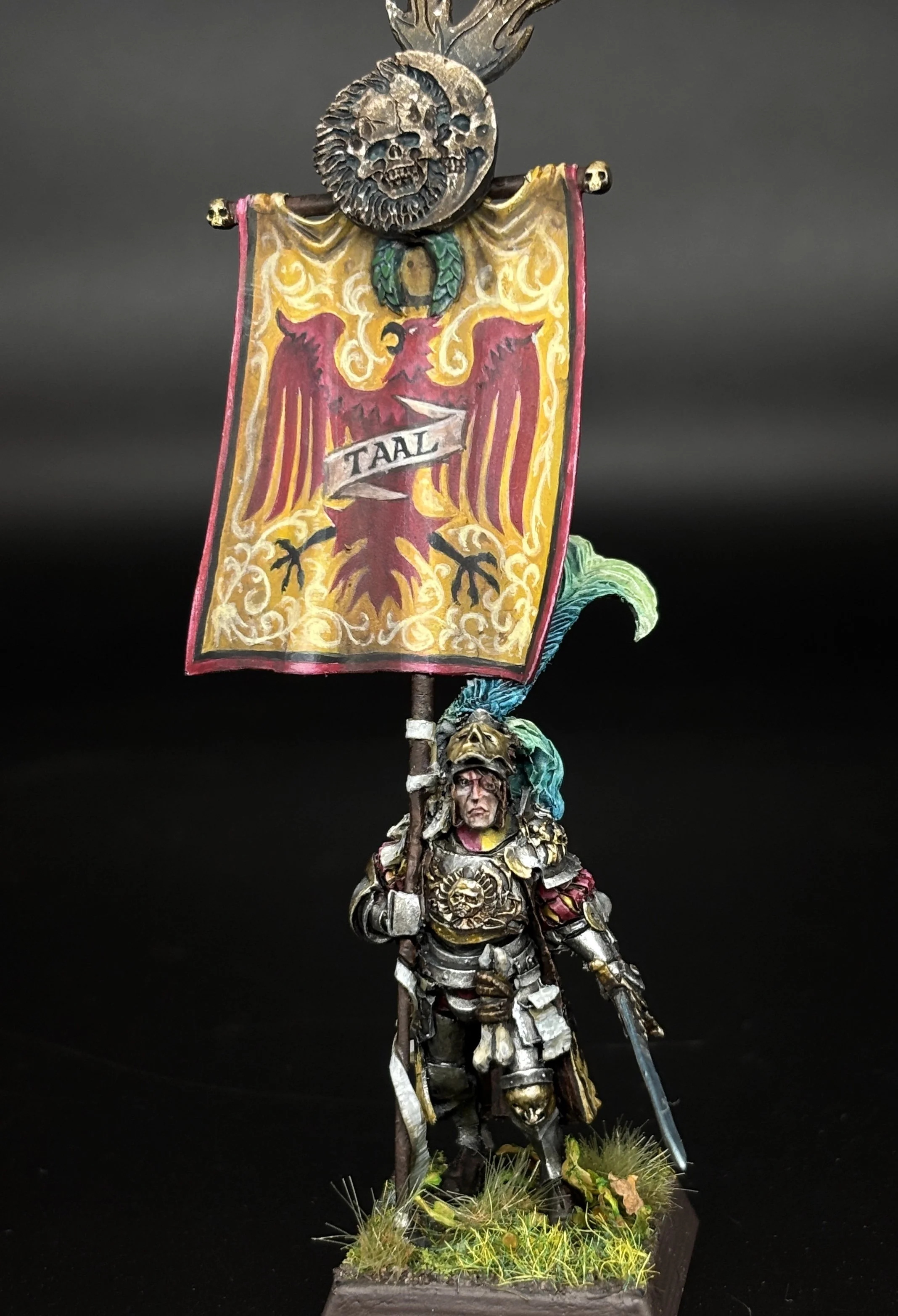 A detailed miniature figurine of a medieval knight dressed in armor, holding a flag with a red crest and a banner that reads 'Taal'. The figure is mounted on a base with grass and small plants, and features decorative skull motifs on the armor and banner.