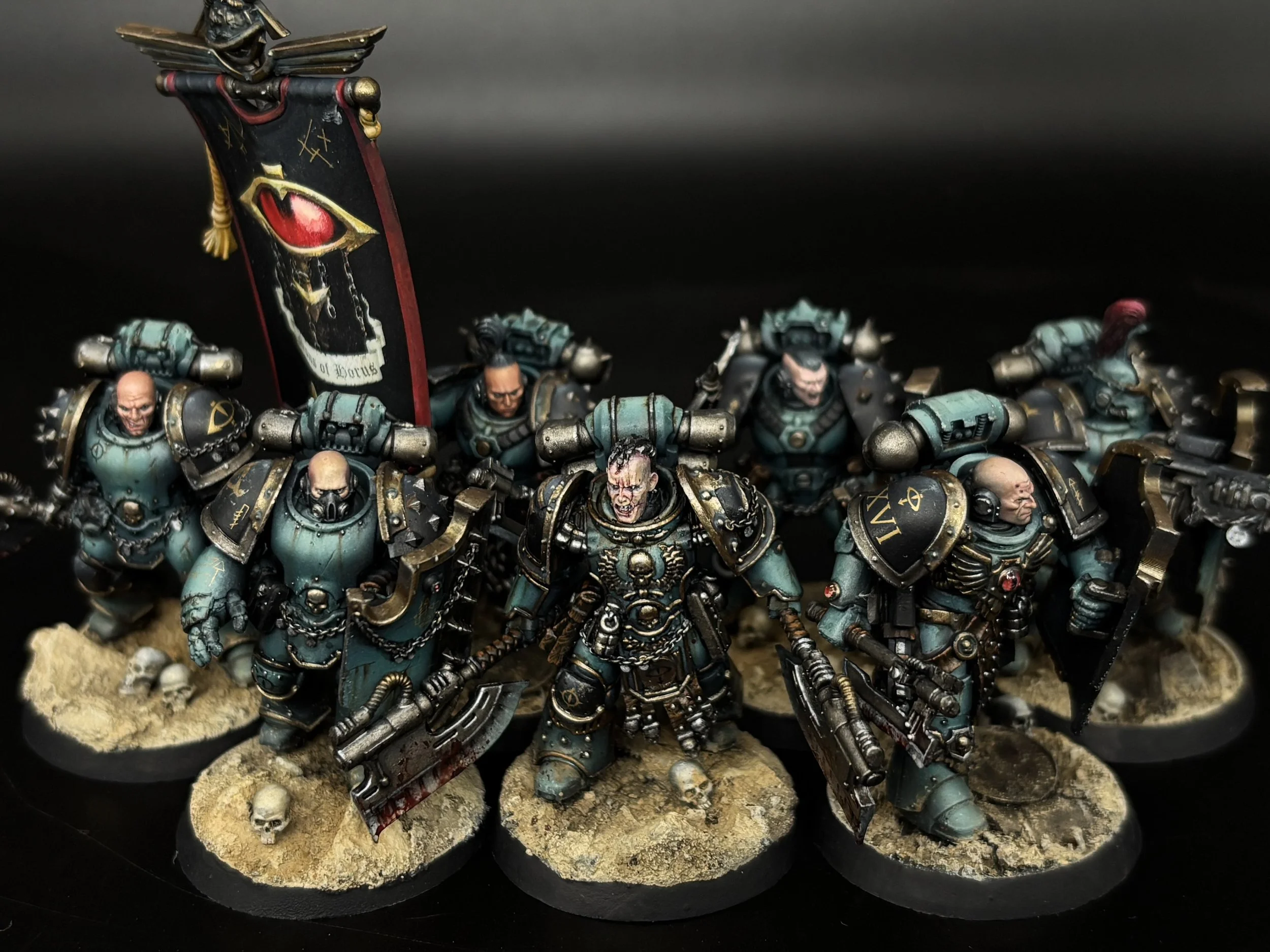 Miniature wargaming tabletop scene featuring armored soldiers with a banner depicting an eye and blood drops, positioned on a textured sandy base with skulls and bones.