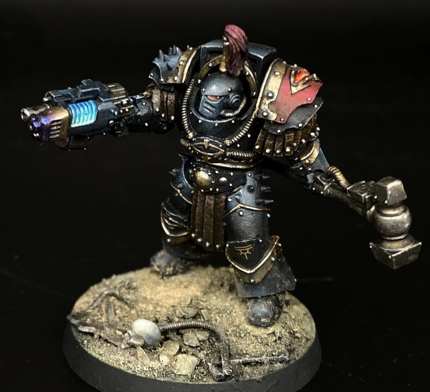 Miniature figurine of a heavily armored space marine wielding a gun and a hammer, standing on a textured base with debris and skulls, against a black background.