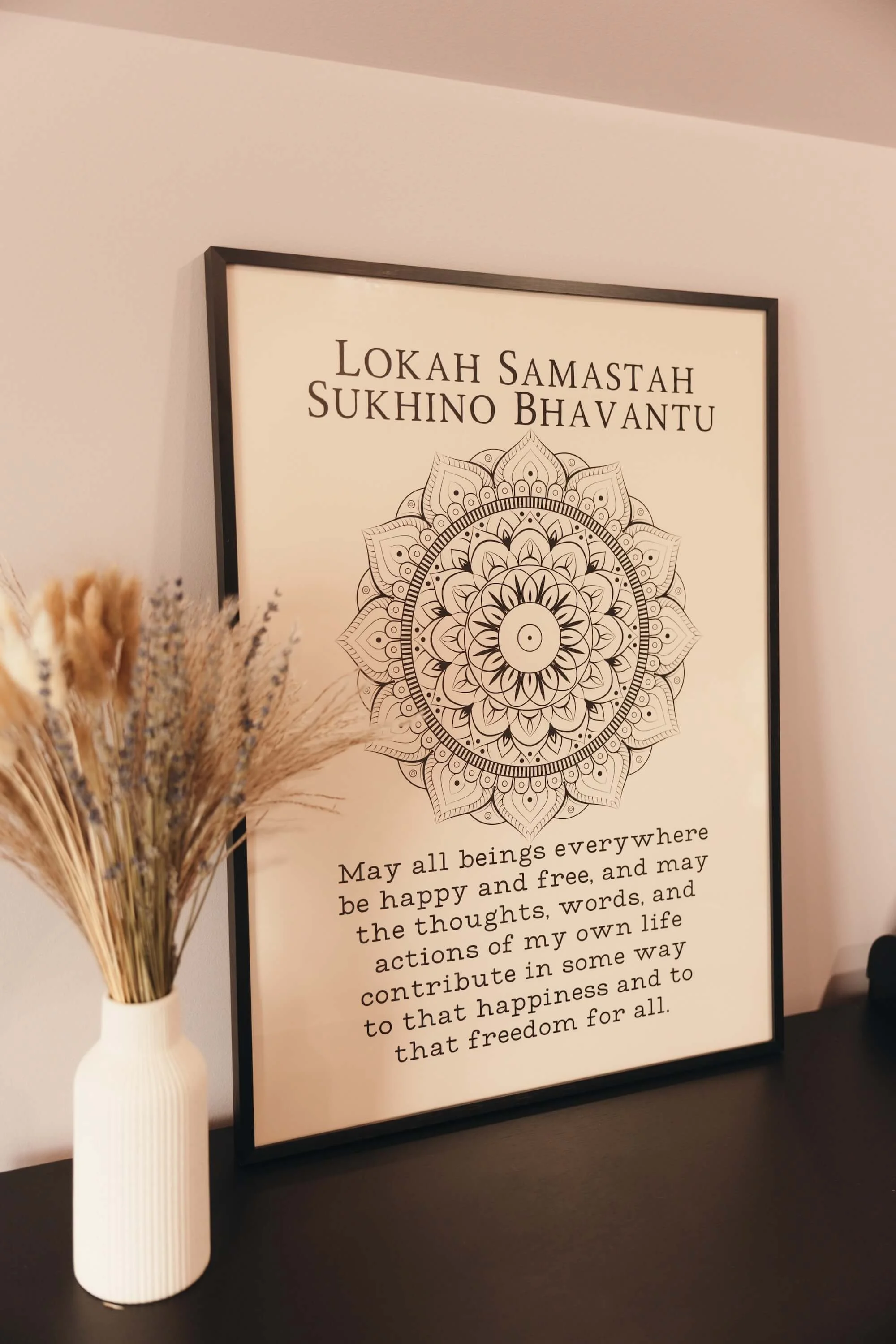 A framed poster with a mandala design and text about happiness and freedom, placed on a surface next to a white vase with dried flowers.