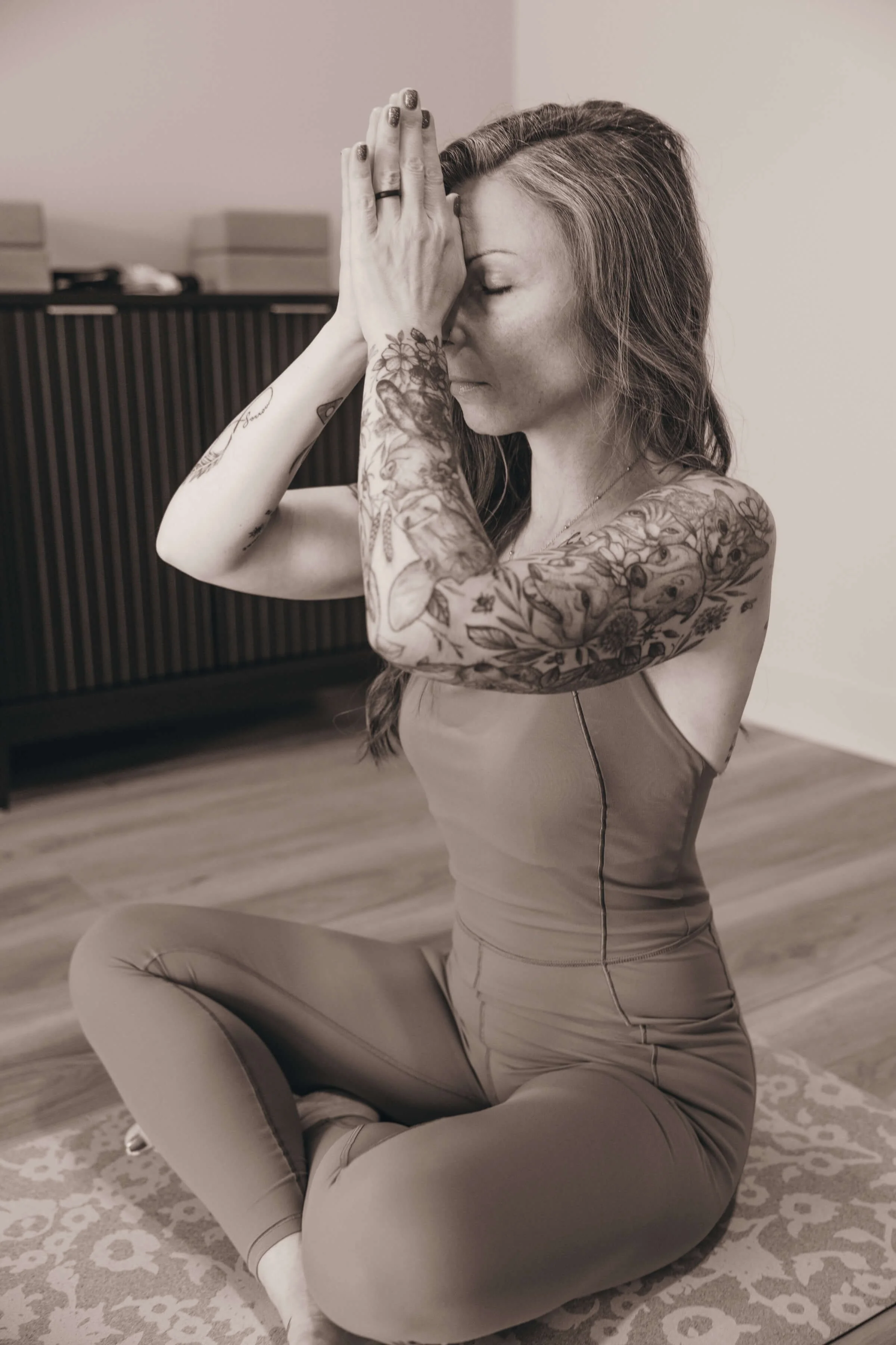 A woman with long hair, tattoos on her arms, and a black ring on her finger, sitting on a patterned rug with her eyes closed, hands pressed together near her face, practicing yoga or meditation in a room with a wooden floor and a cabinet in the background.
