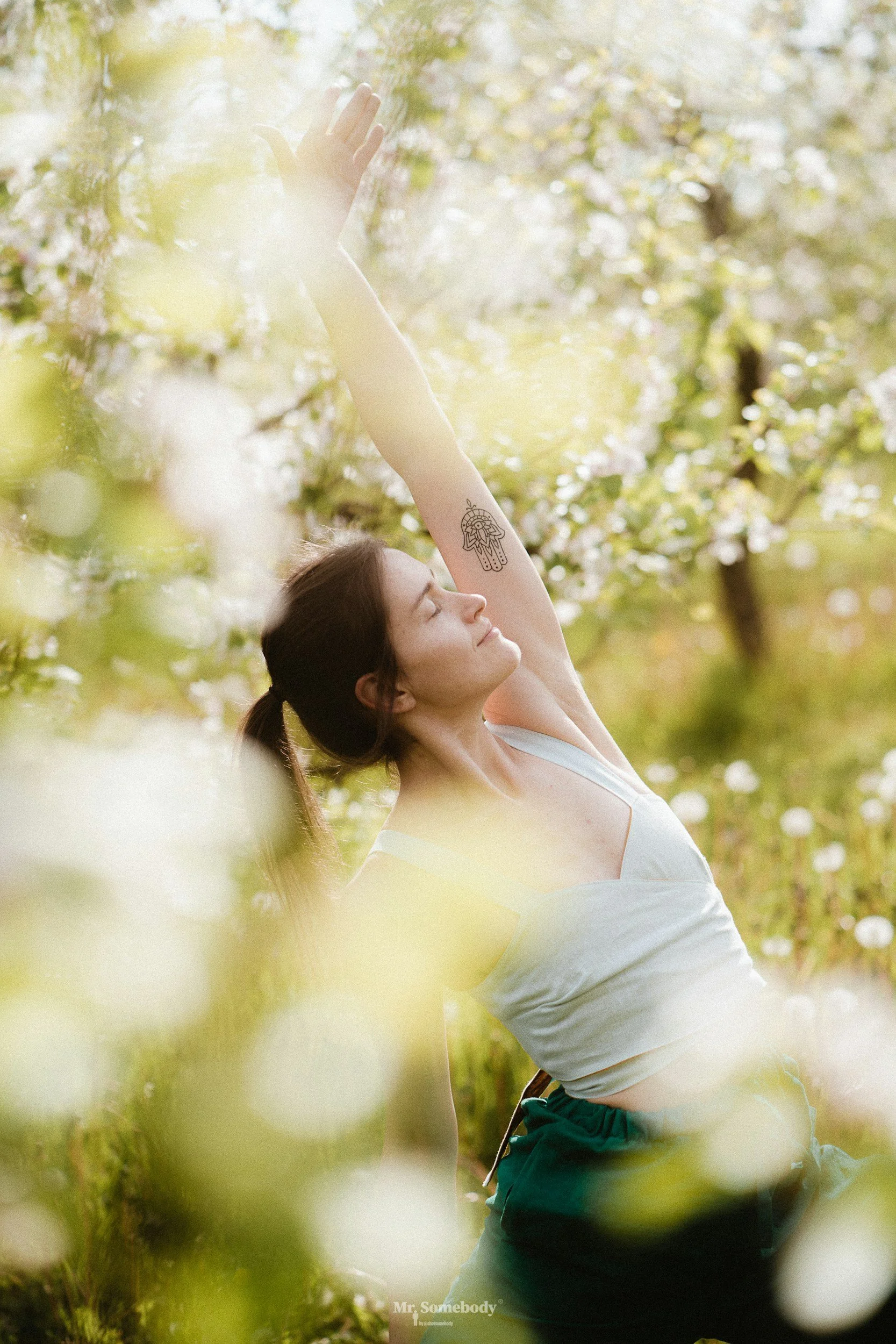 A woman with a tattoo on her arm is doing yoga outdoors among flowering trees on a sunny day.